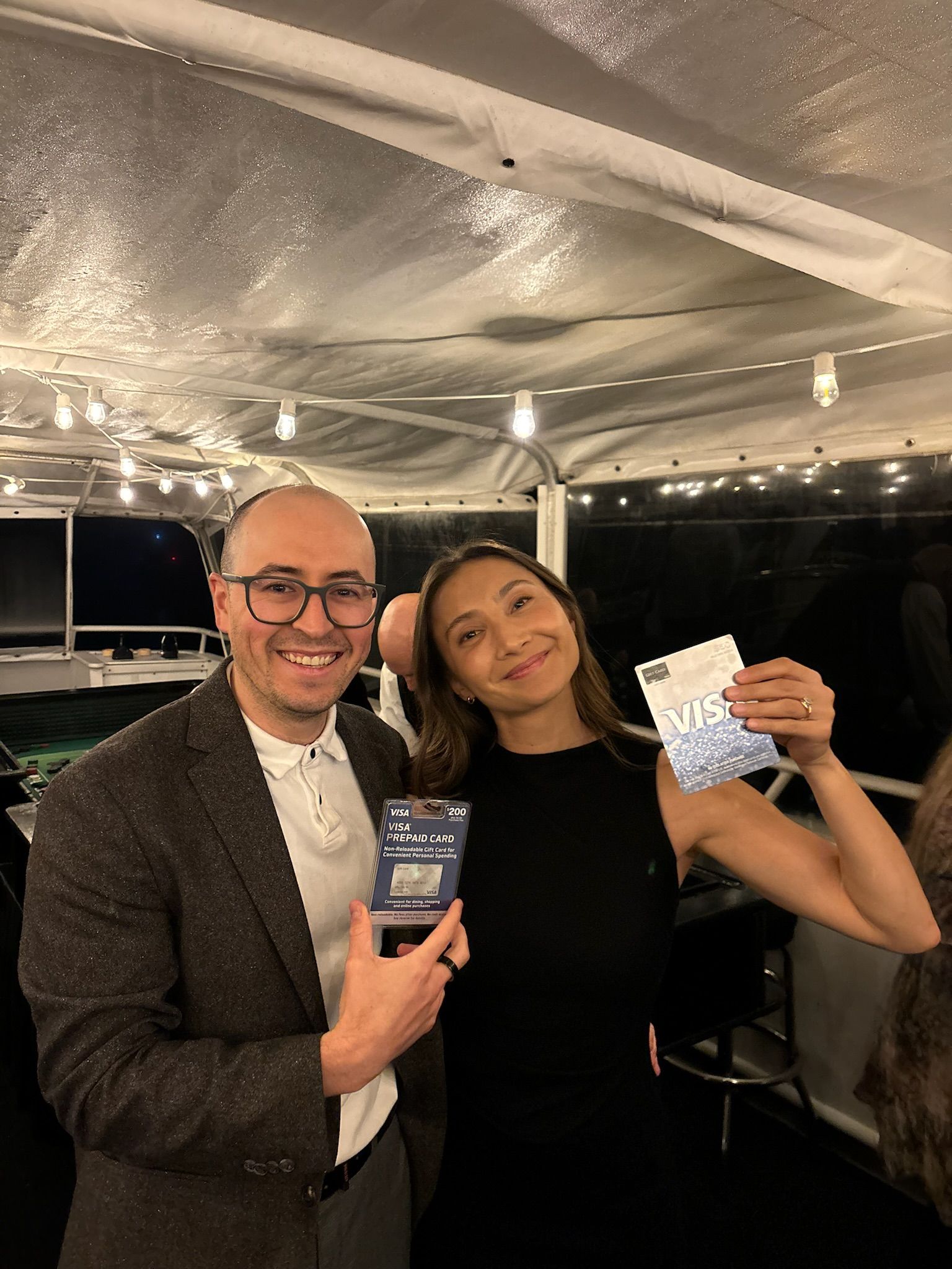 Two people smiling indoors, holding cards under string lights at a party.