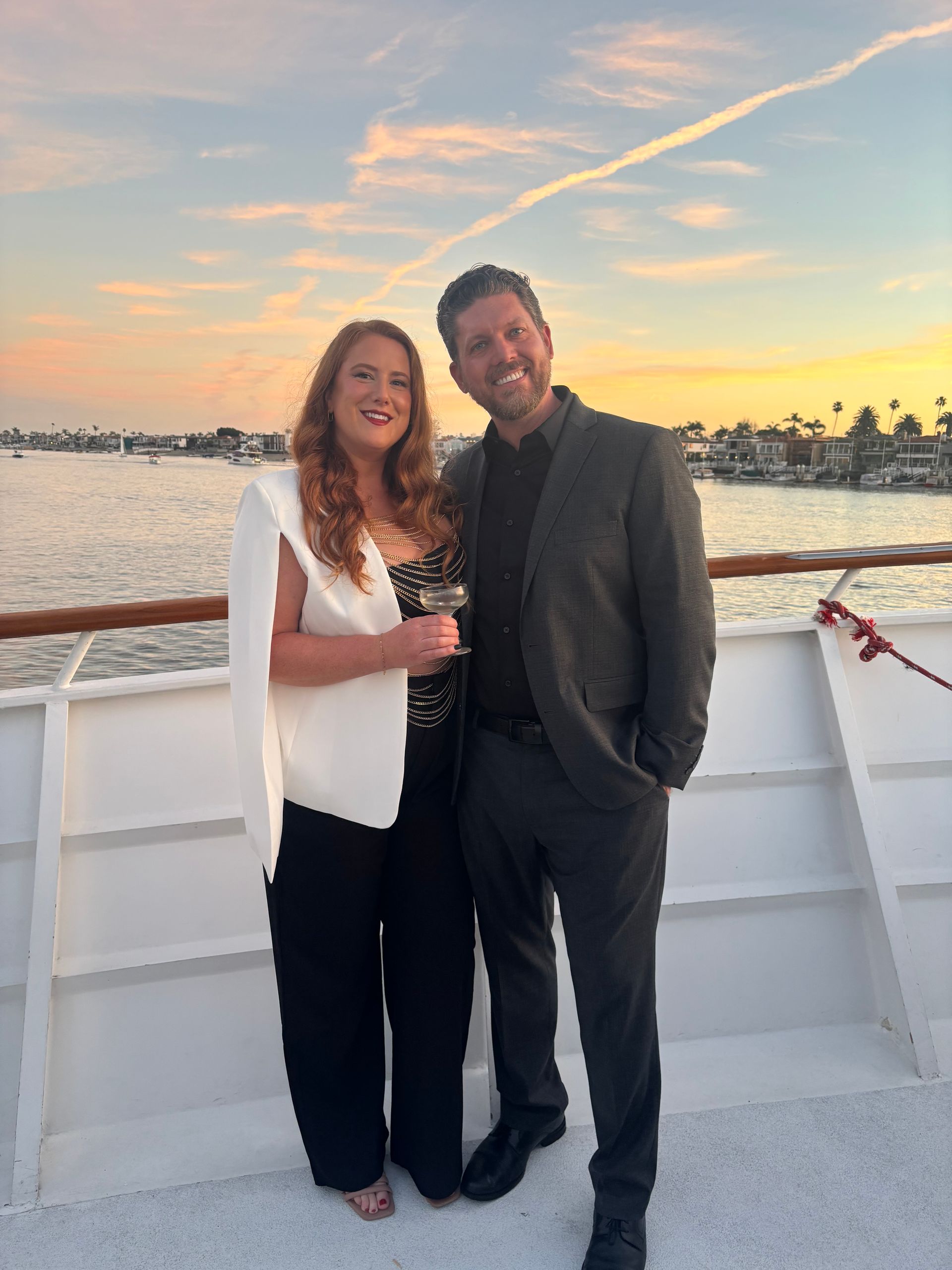 Two people standing on a boat deck at sunset, smiling with water and shoreline behind them