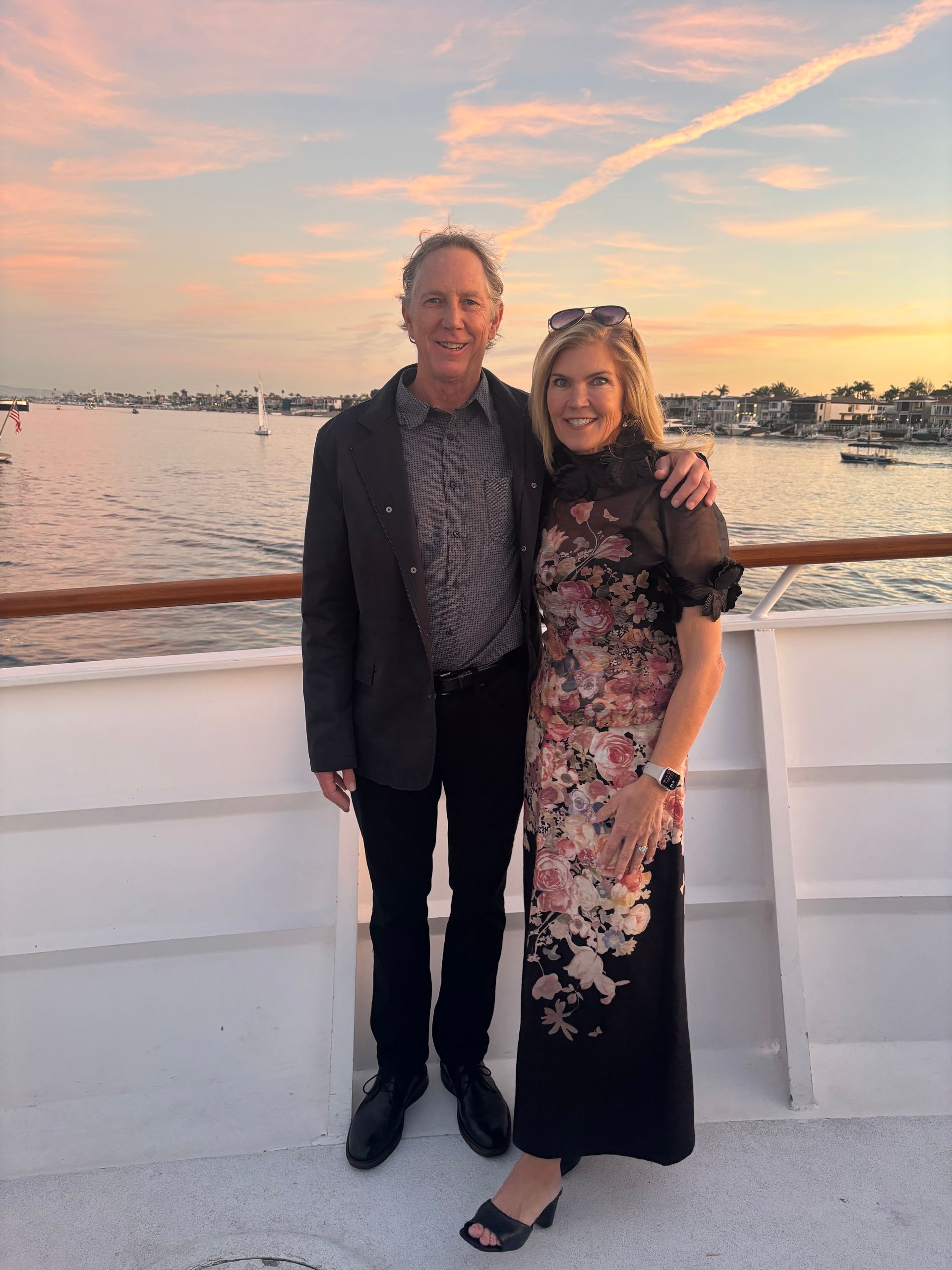 Two people stand on a boat deck at sunset, smiling with a calm waterfront in the background.