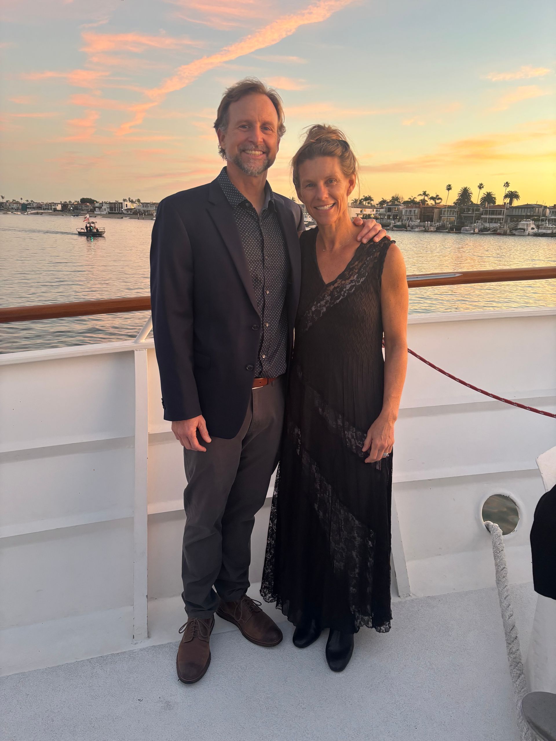 Two people in formal wear standing on a boat deck at sunset by the water.