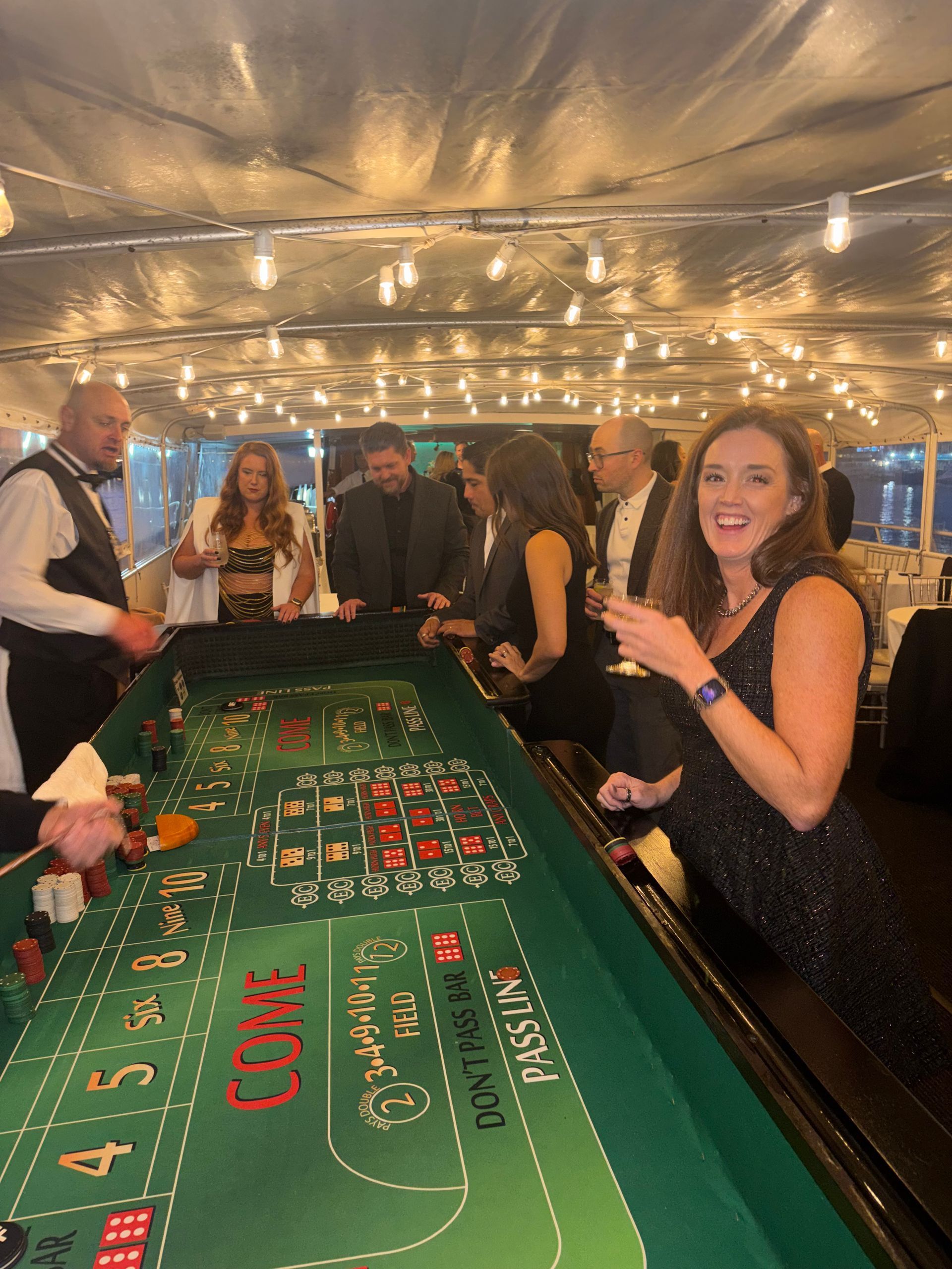 People playing roulette at a casino table under warm string lights, with a woman smiling in the foreground