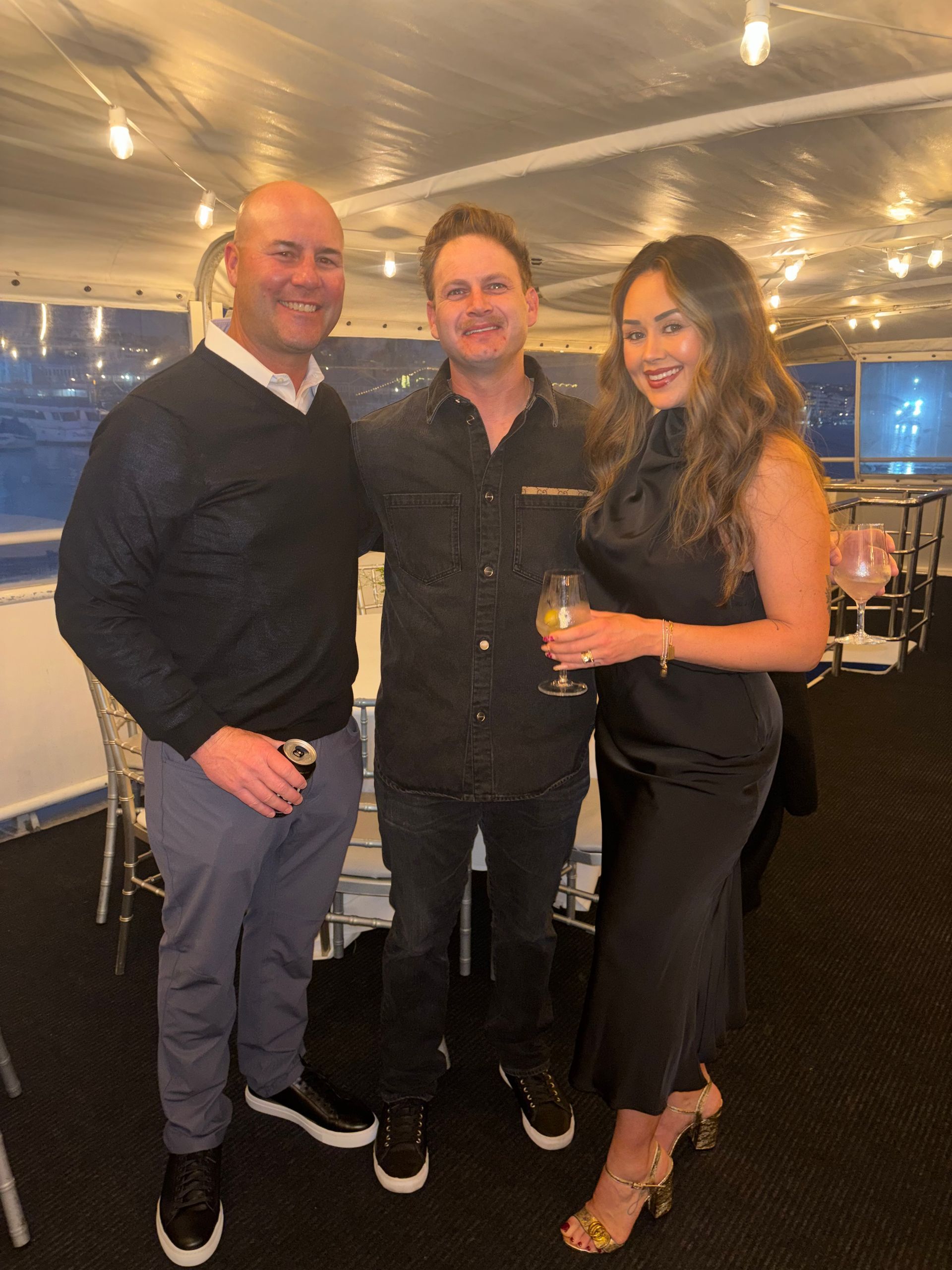 Three people smiling and holding drinks at an indoor evening gathering with warm string lights.