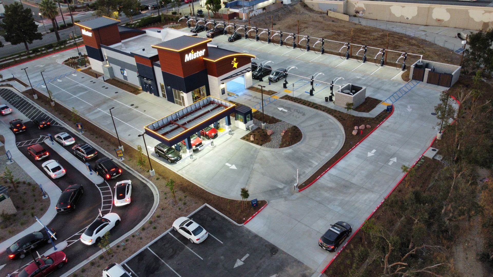 Aerial view of a small modern gas station with a lit canopy, parking lot, and surrounding streets at dusk