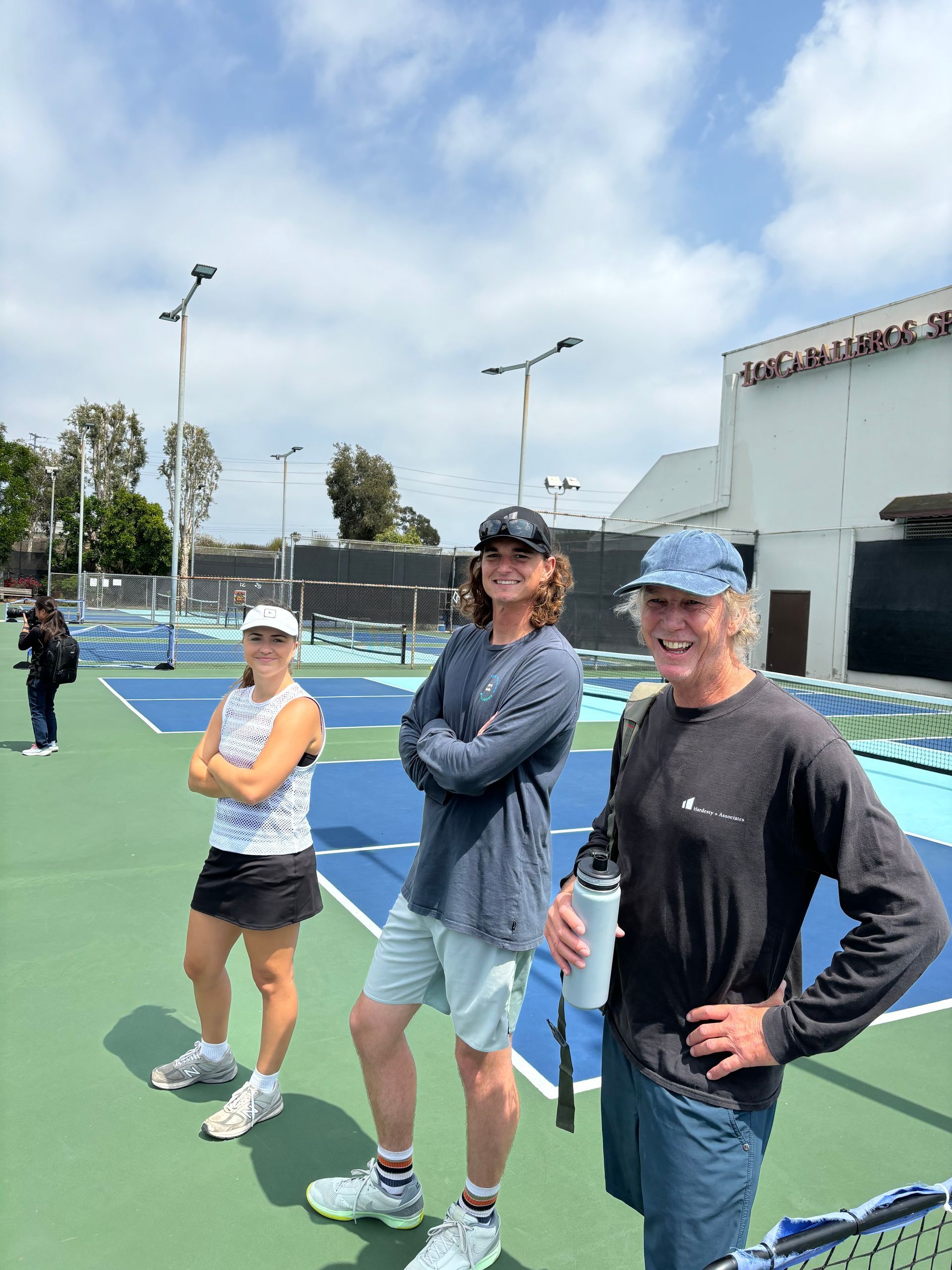 Three people standing on a tennis court, two smiling at the camera under a bright blue sky.