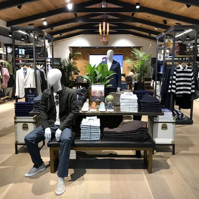 Clothing store display with mannequins, folded shirts, jackets, and potted plants under exposed wooden beams