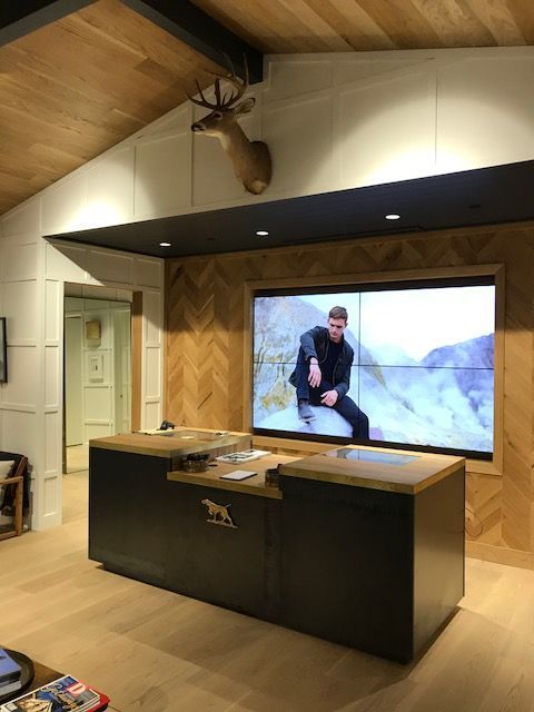Modern kitchen showroom with black island, wood ceiling, and wall screen showing a person outdoors