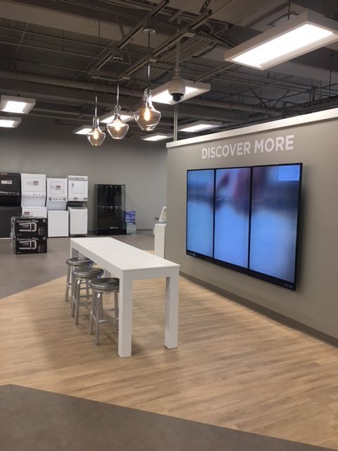 Modern showroom with display screens, white table, stools, and “DISCOVER MORE” wall sign
