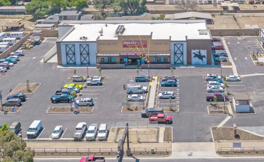 Aerial view of a shopping center parking lot with many parked cars and storefronts at the entrance.
