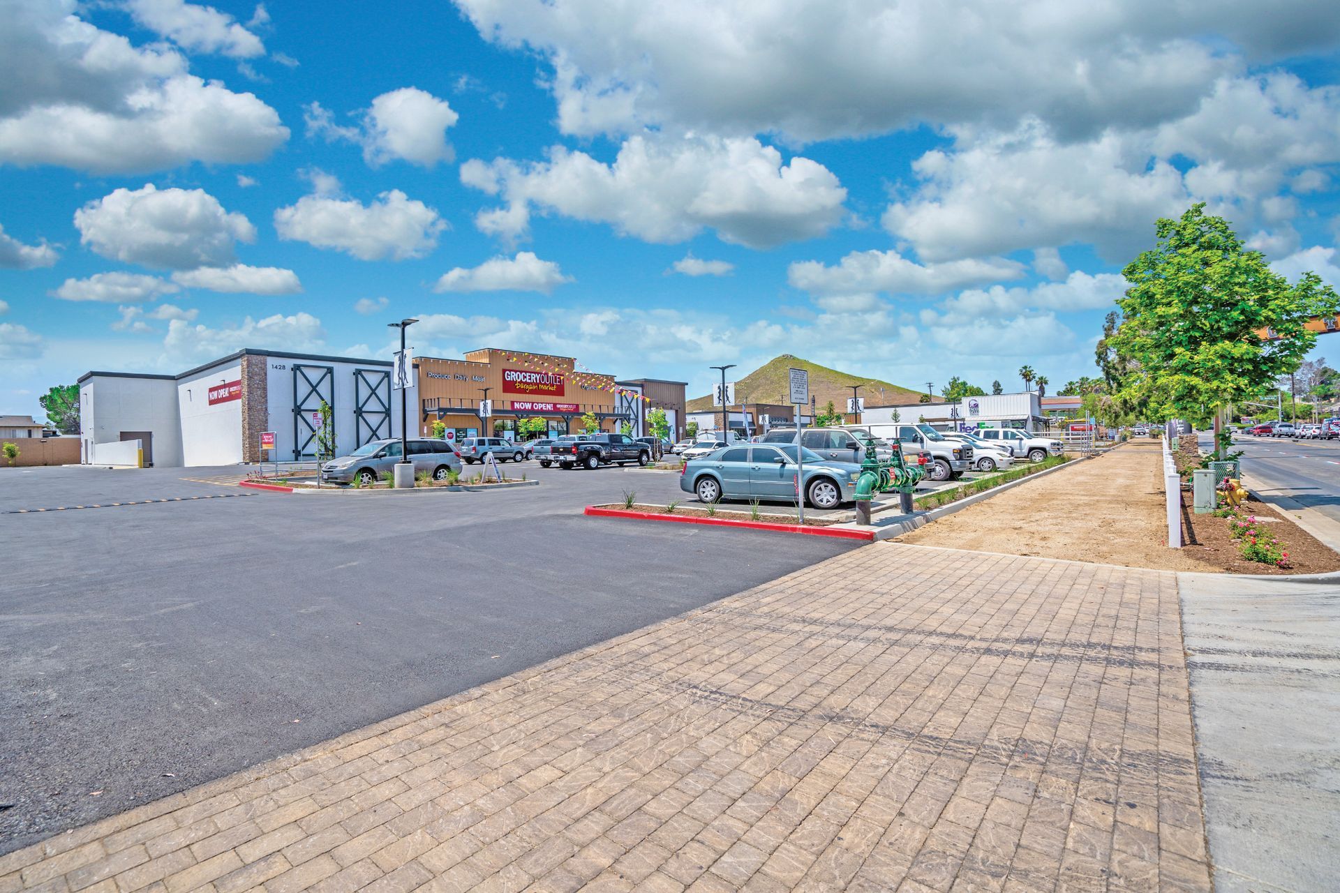 Wide parking lot beside storefronts under a bright blue sky with clouds, cars parked along the curb.