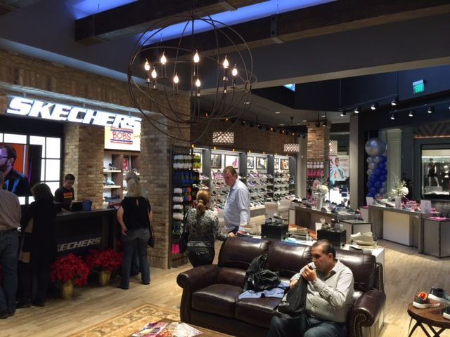 Inside a Skechers store, shoppers browse displays under warm pendant lights, with a man seated on a sofa in front.