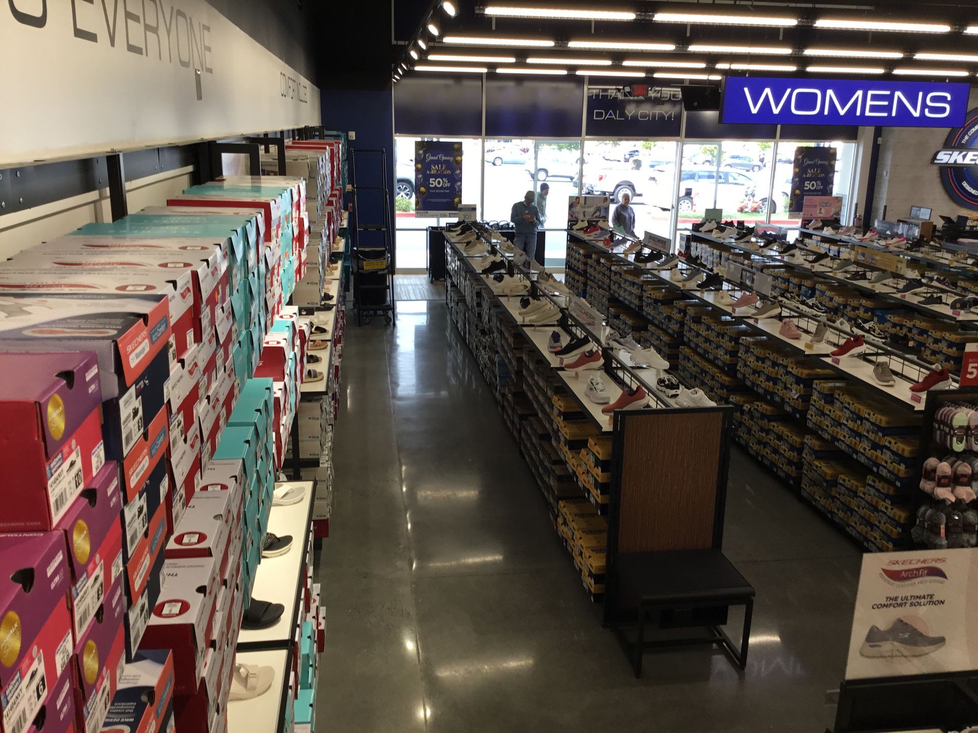 Shoe store aisle with rows of boxes and shelves, including a neon “WOMENS” sign near the entrance