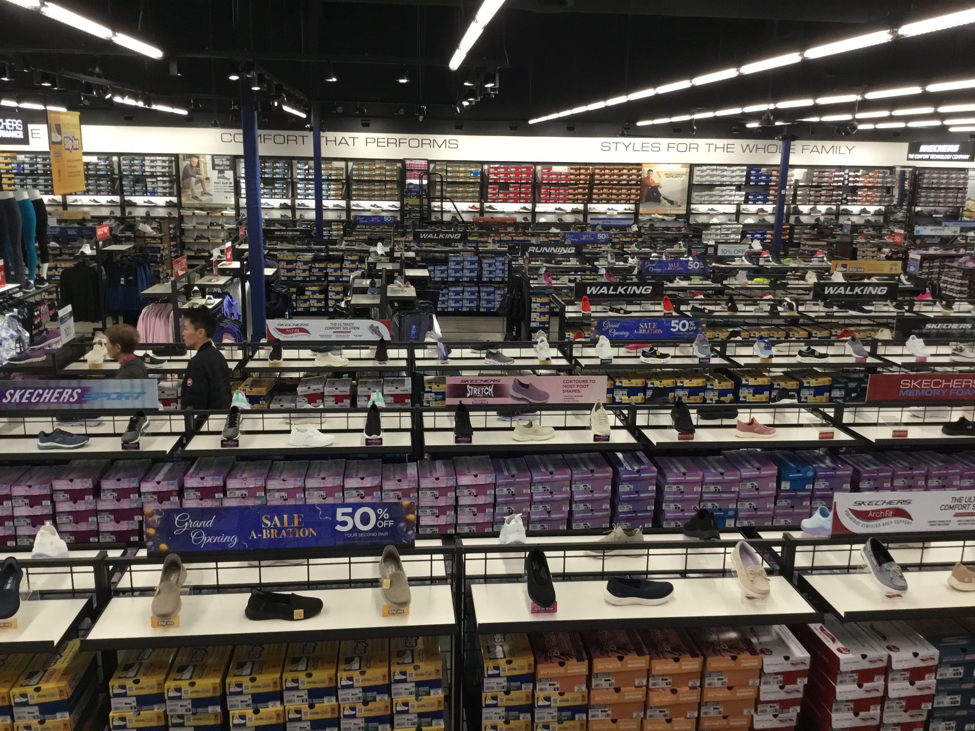 Wide view of a bookstore aisle with shelves of books, signs, and a few shoppers browsing