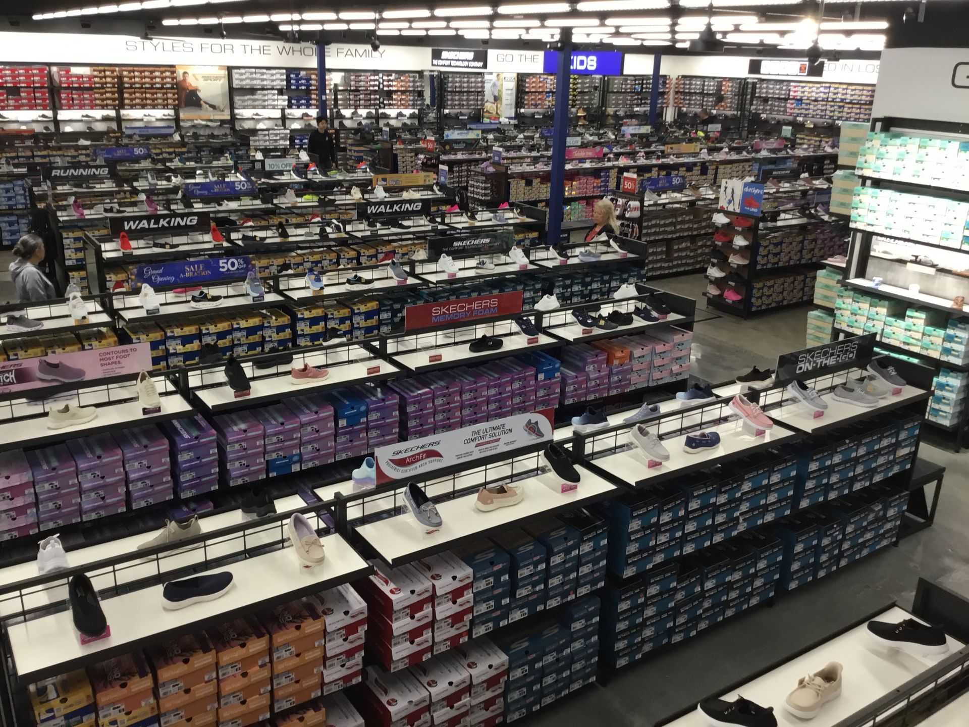 Retail store aisle with rows of shoes and boxes on display shelves under bright overhead lighting