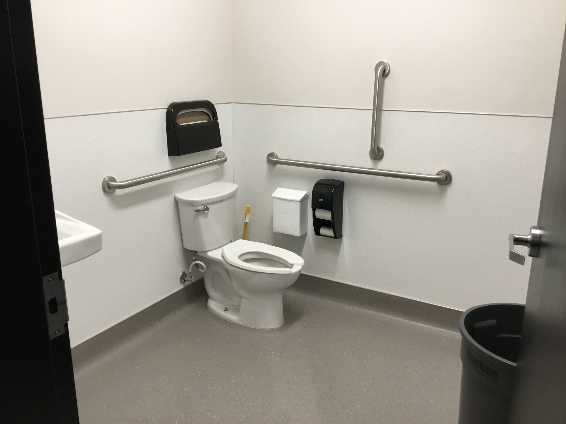 Accessible restroom with toilet, grab bars, wall-mounted dispensers, and a trash can