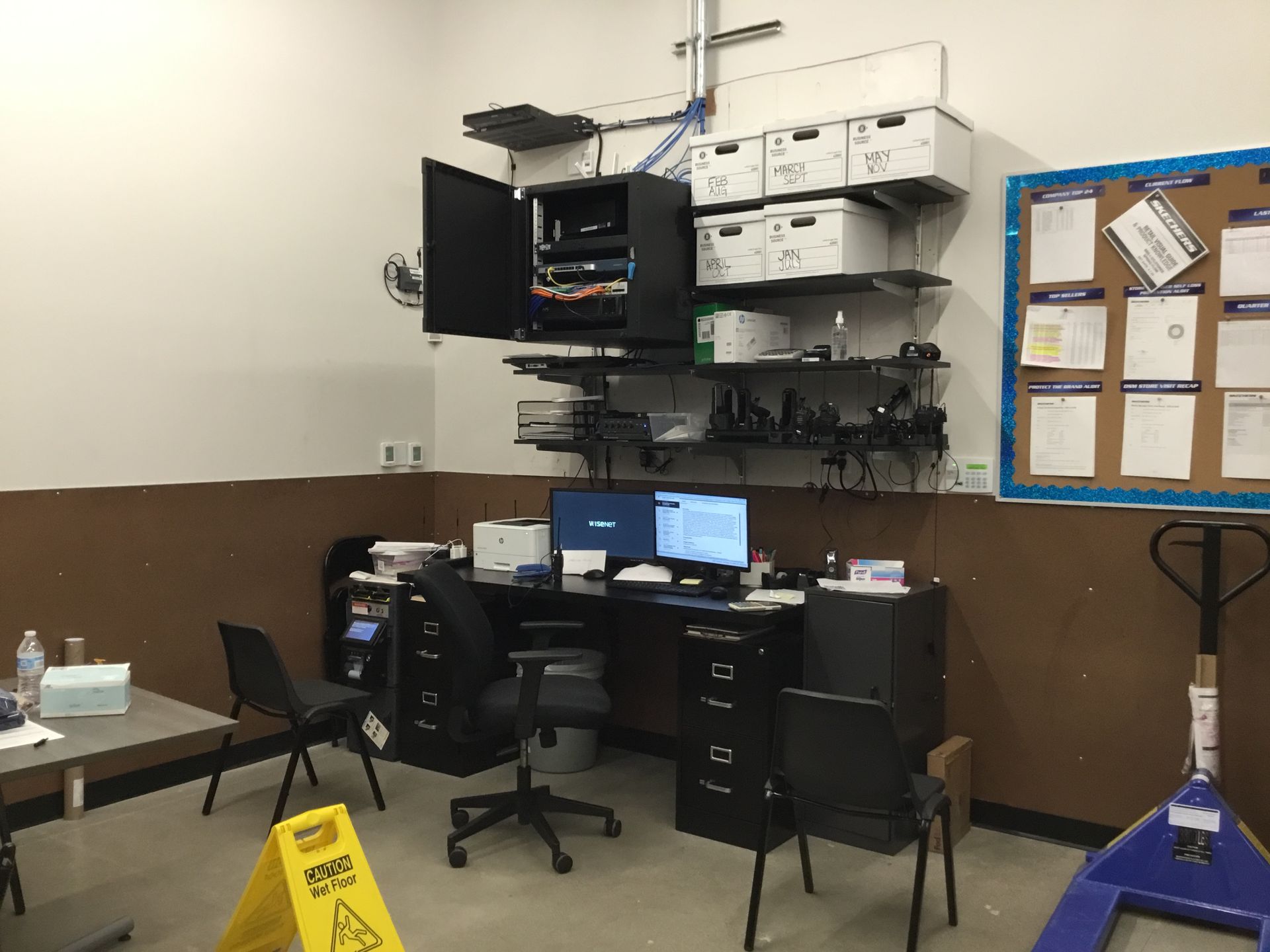 Small office workstation with dual monitors, shelves of equipment, and a yellow wet floor sign.