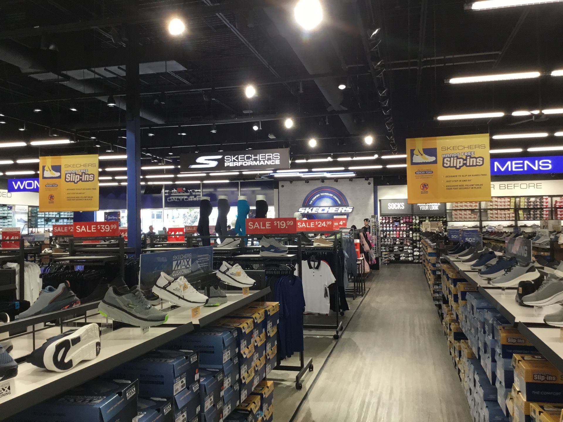 Shoe store aisle with rows of sneakers on display and sale signs overhead under bright ceiling lights