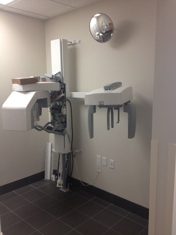 Dental exam room with X-ray machine, wall-mounted sink, and round mirror on beige walls.