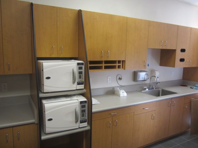 Small kitchen with wood cabinets, two stacked white microwaves, sink, and countertop appliances.