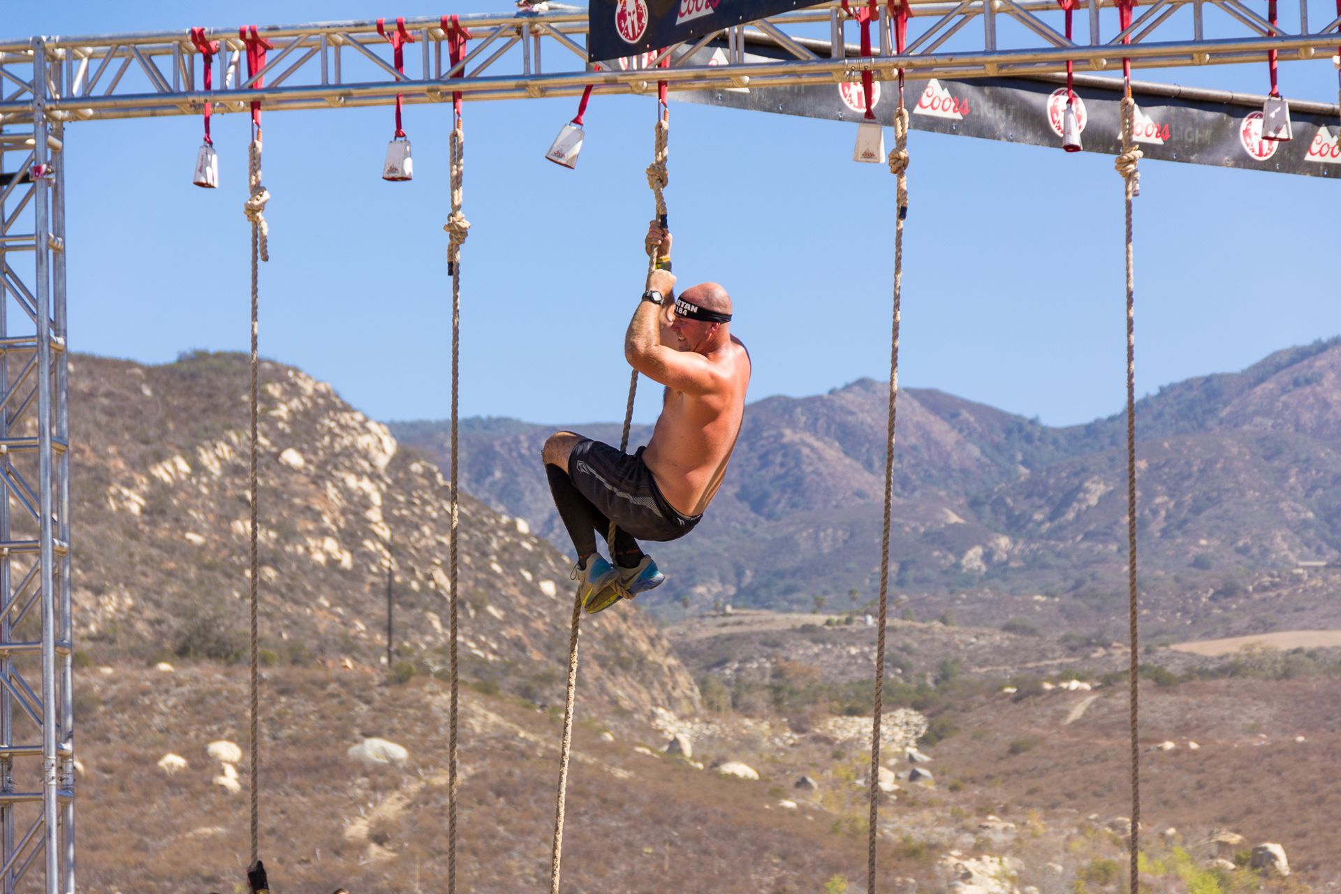 A shirtless climber hangs from ropes on an outdoor obstacle course with mountains in the background.