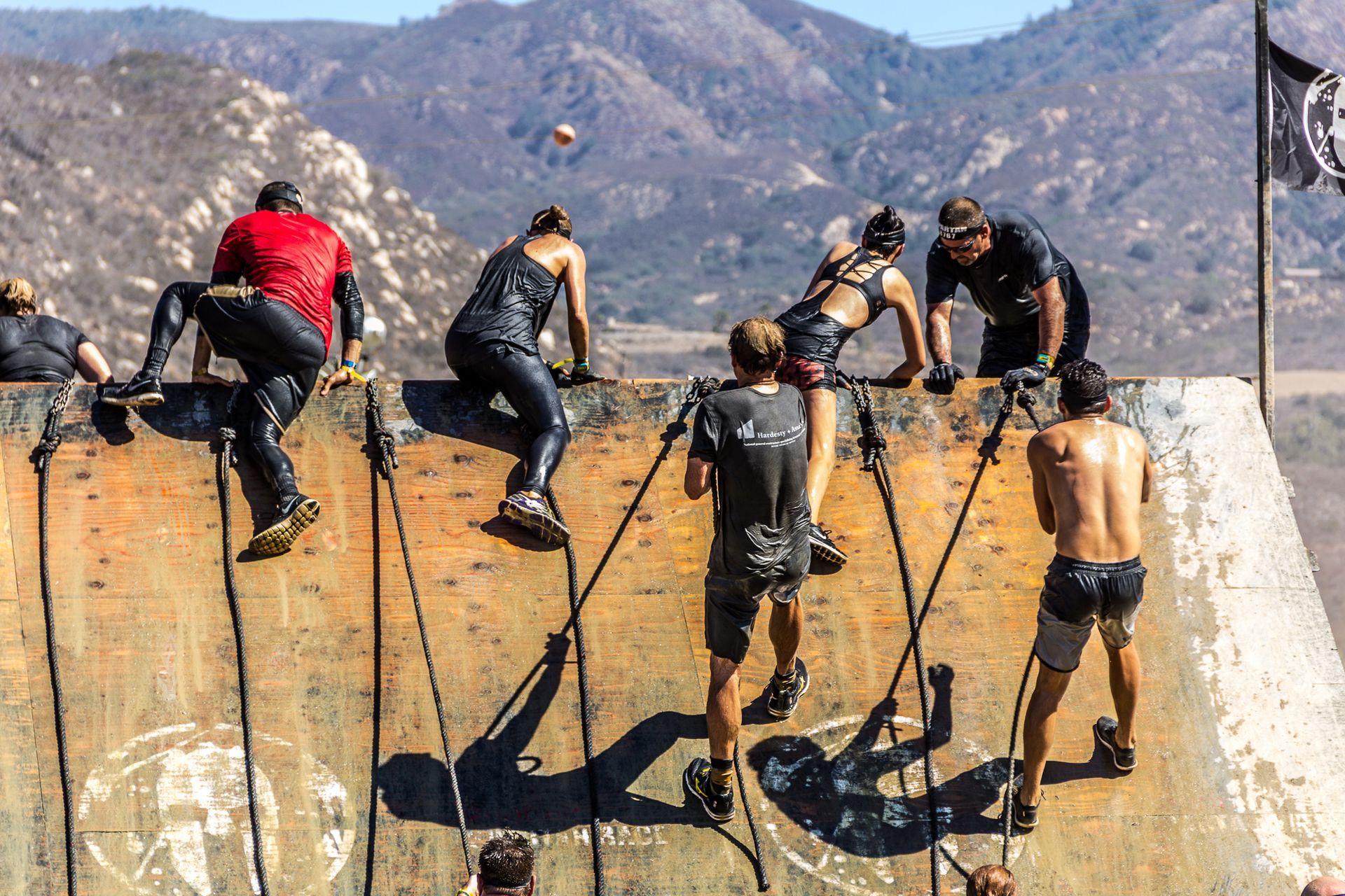 People climbing over a muddy obstacle wall with ropes, mountains in the background