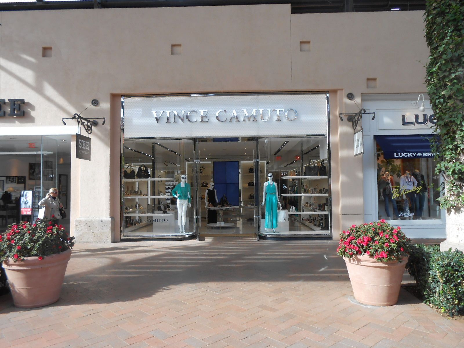 Storefront entrance with mannequins and clothing display, flanked by large potted flowers on a sunny walkway