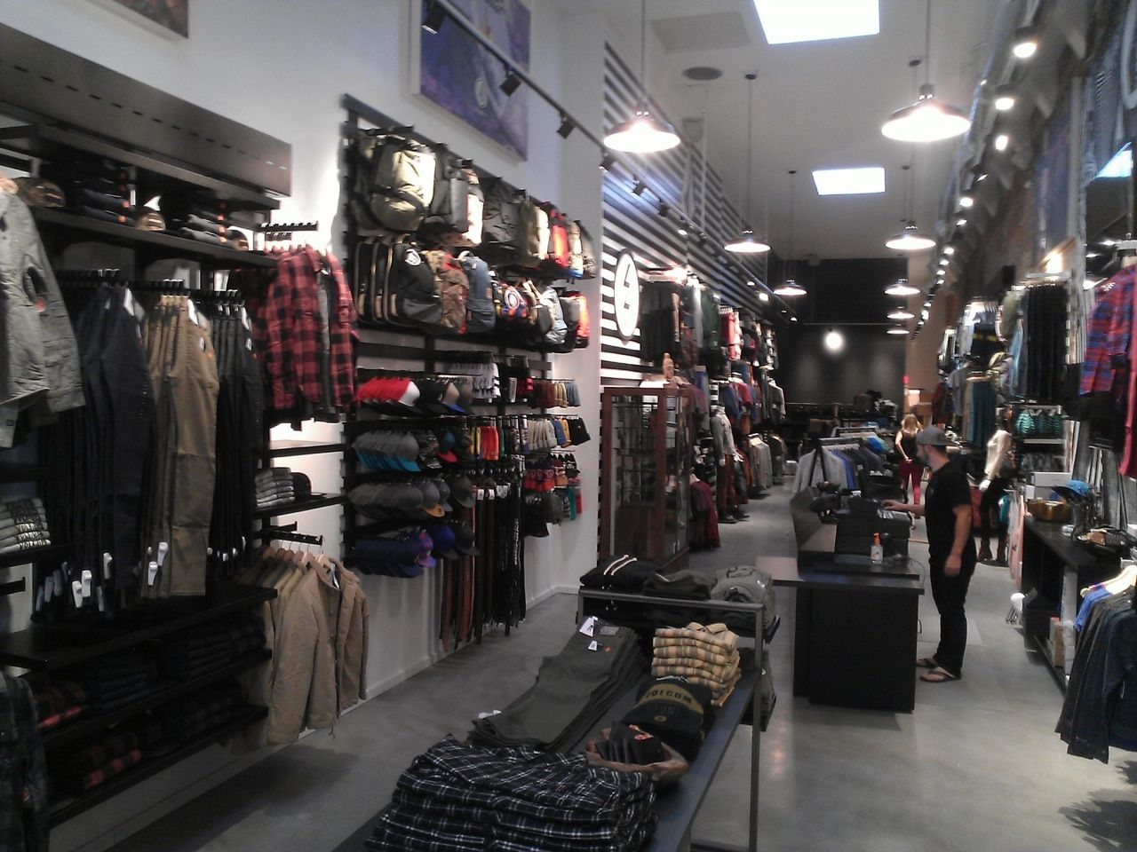 Clothing store aisle with racks of shirts, jackets, and jeans under bright overhead lights