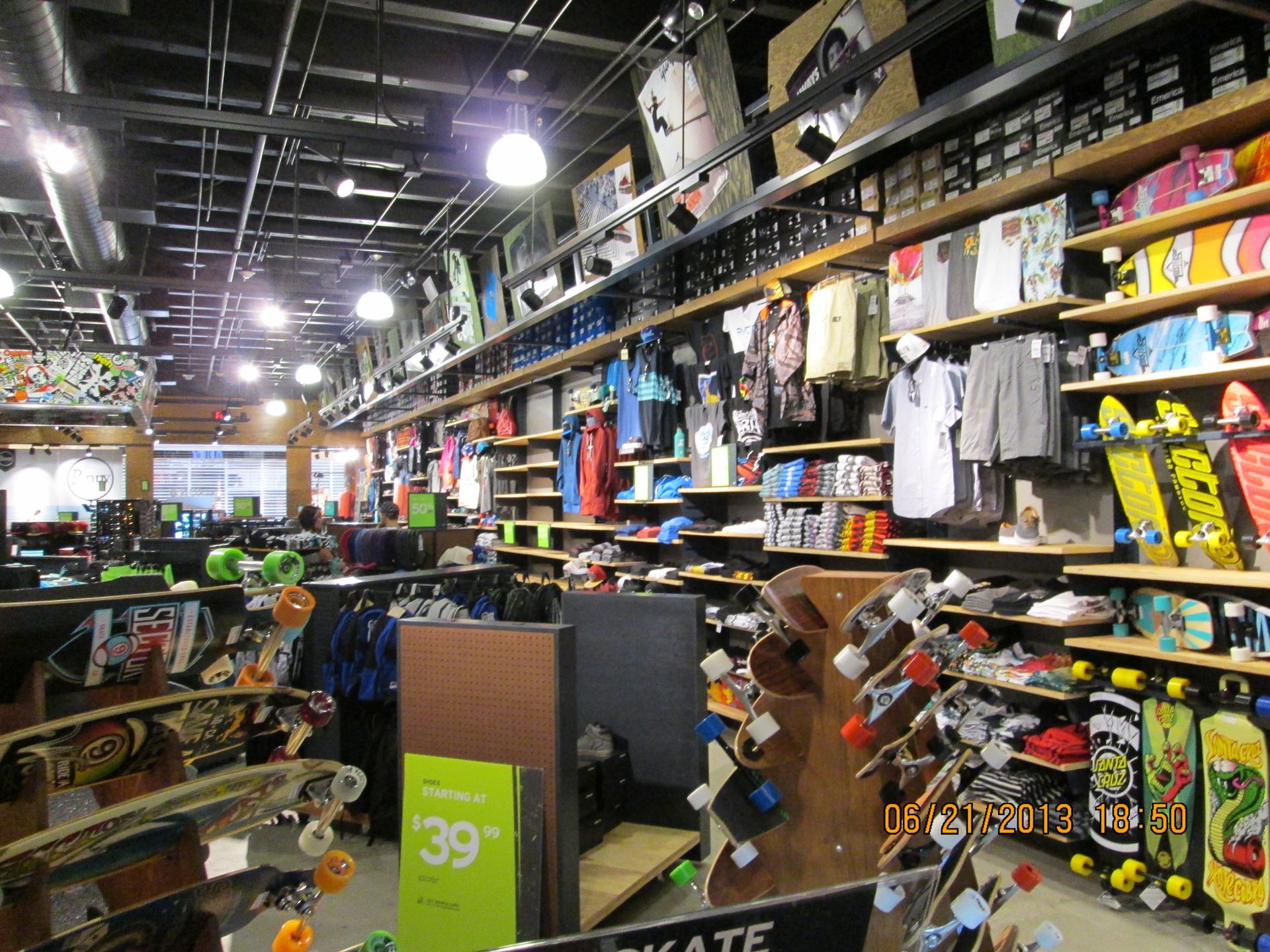 Cluttered retail store aisle with shelves of assorted products and hanging clothes under bright ceiling lights