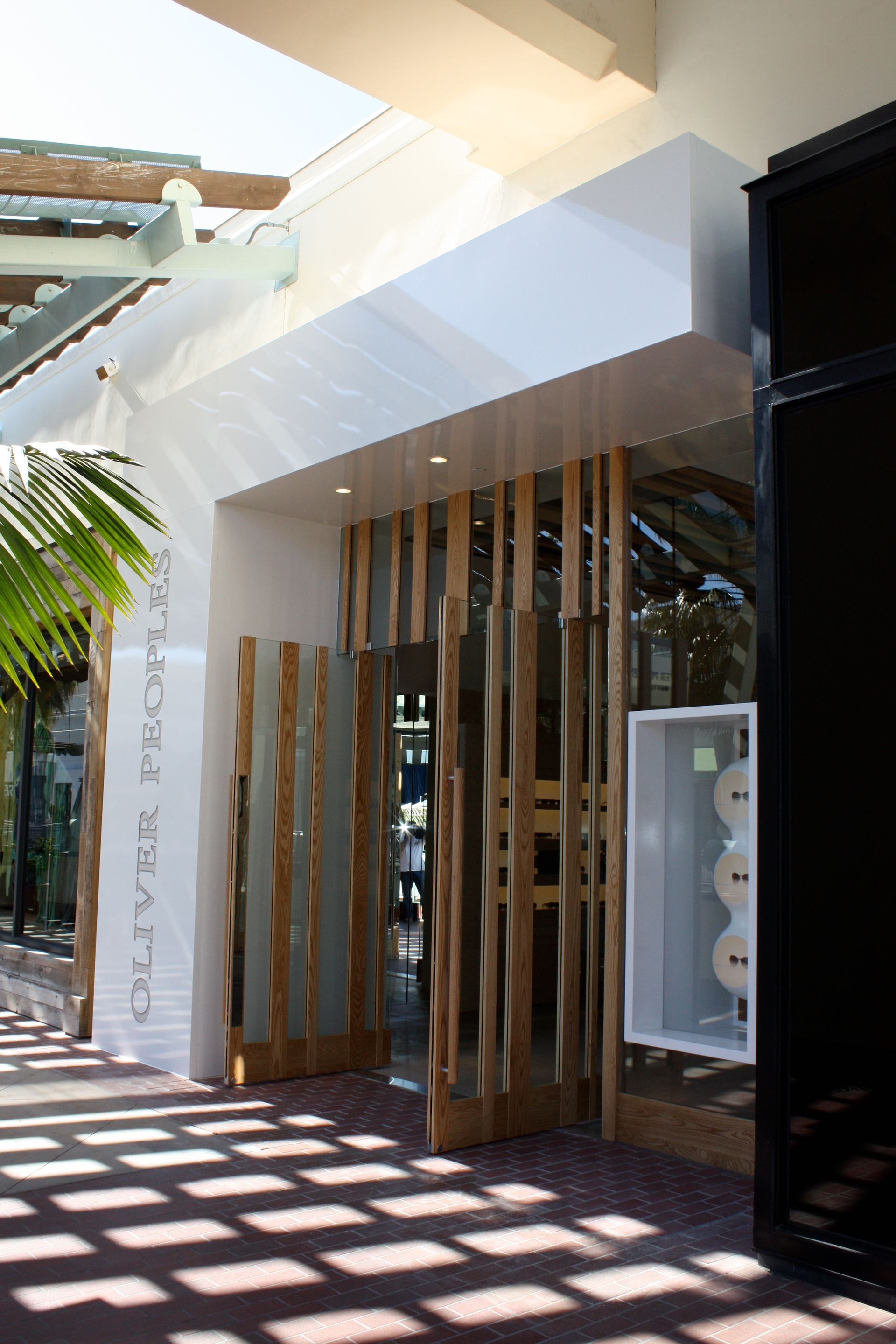 Outdoor storefront with wooden slats, white facade, and sunlit patterned shadows on the walkway
