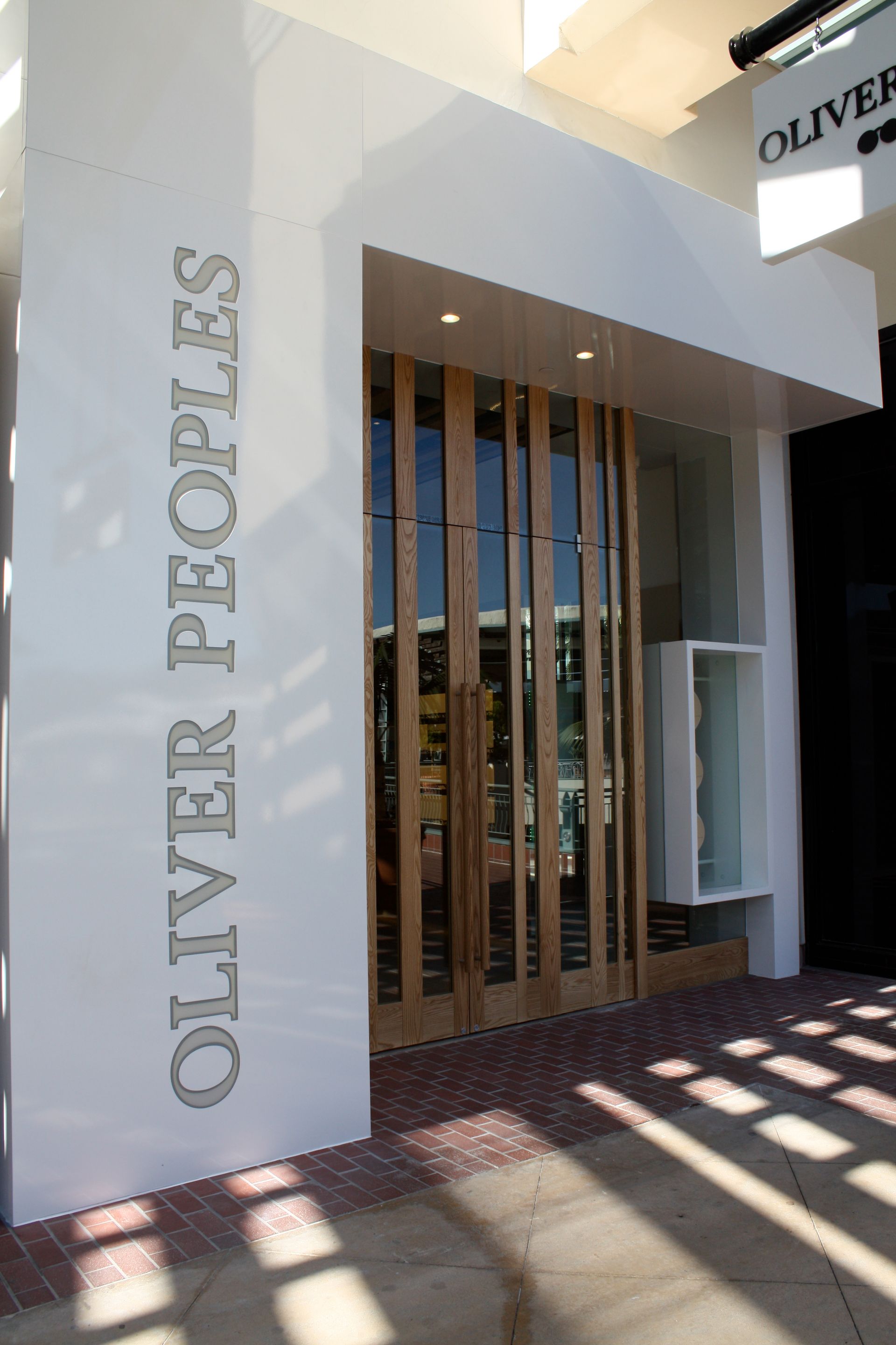 Entrance to Oliver Peoples store with white facade and glass doors casting striped shadows