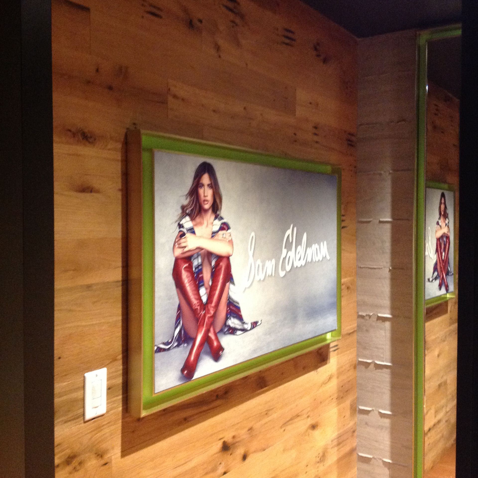 Framed poster on a wood-paneled wall showing a woman seated in red boots, with a mirrored reflection beside it
