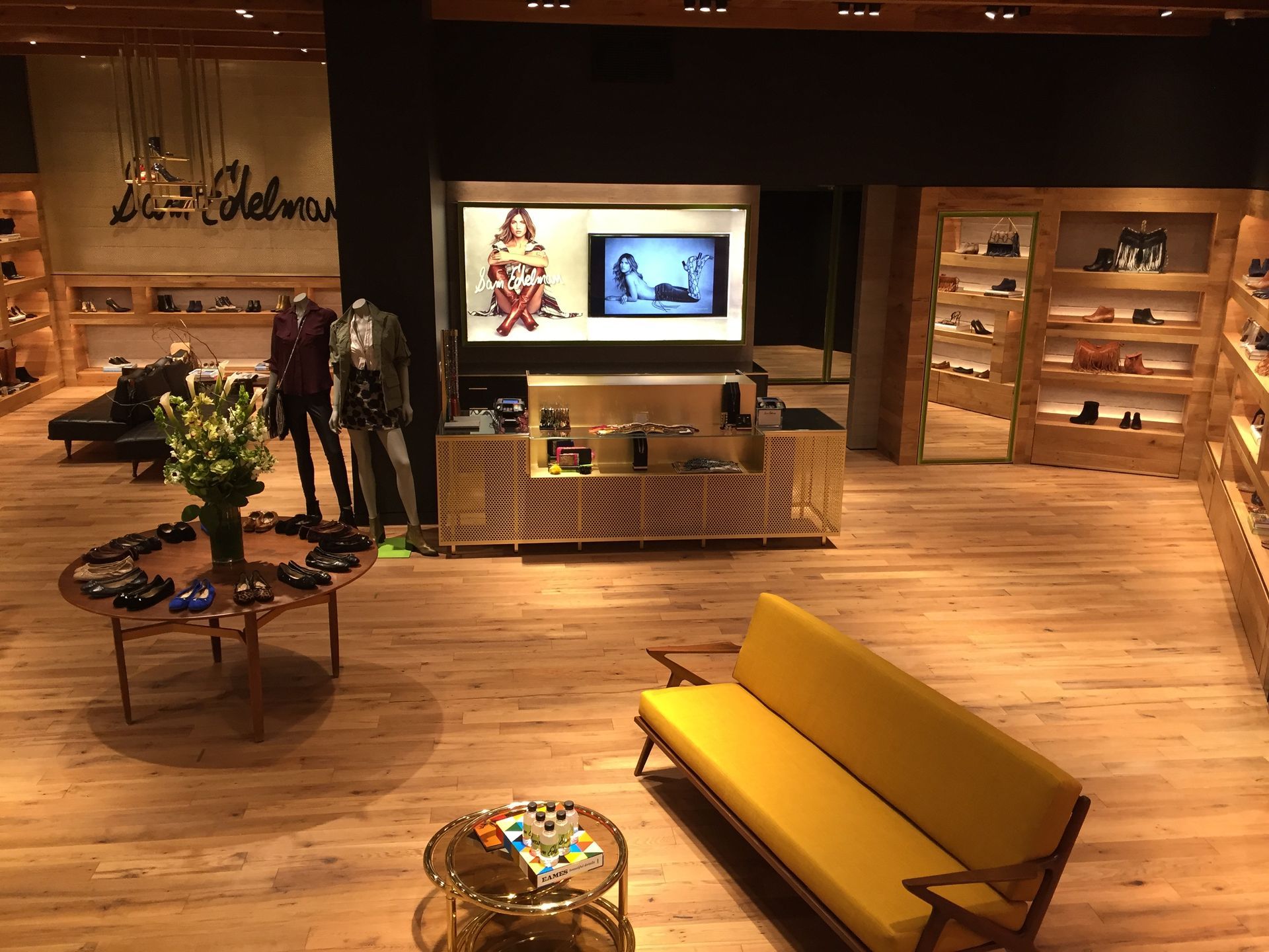 Warm lobby with wood floors, yellow bench, round table, and reception desk under a TV display.