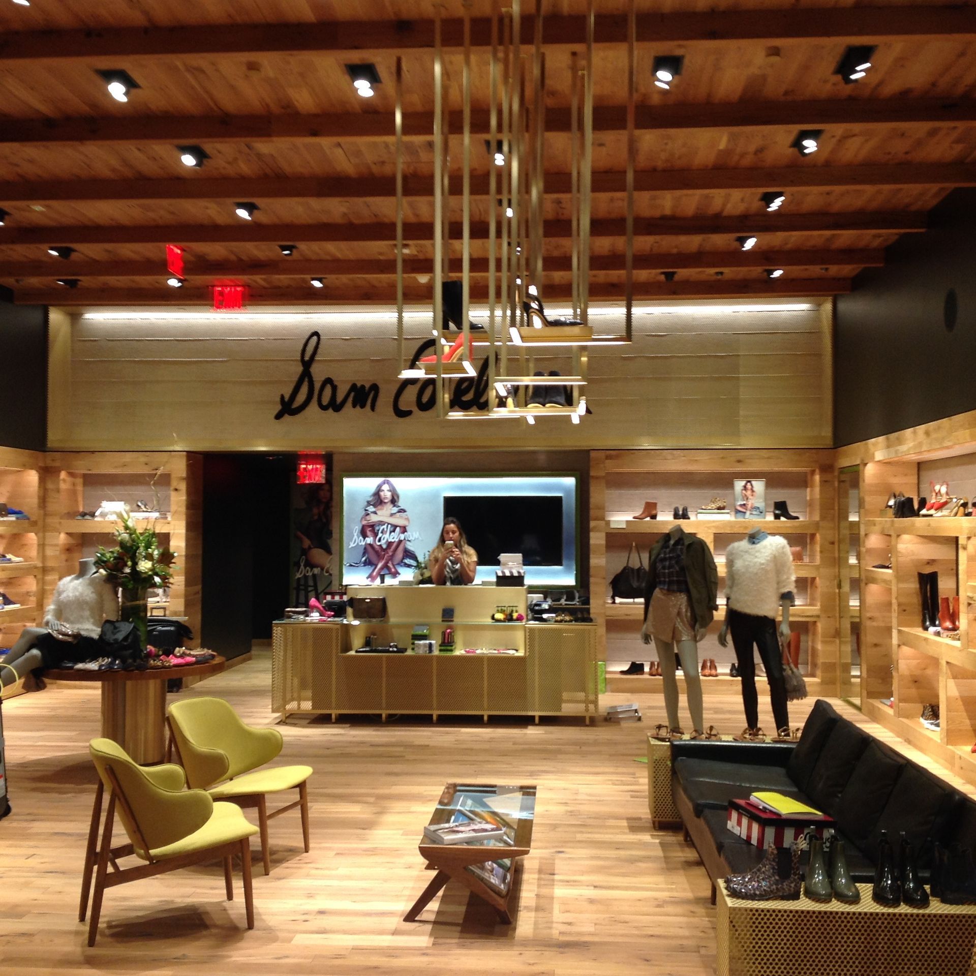 Warm wood-clad clothing store interior with central display table, chairs, and hanging pendant lights