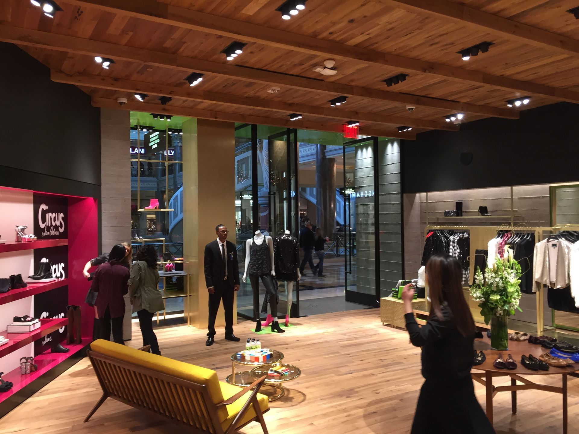 Clothing store interior with mannequins, racks, and shoppers under warm wood ceiling lighting.