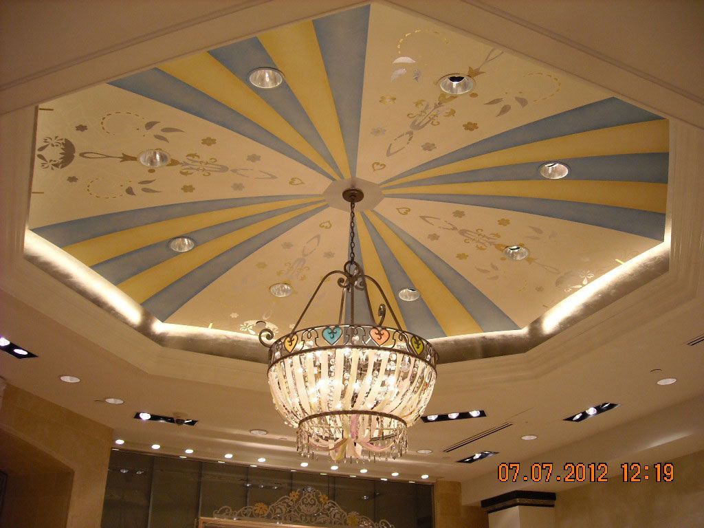 Decorative ceiling with a large crystal chandelier in a bright lobby, in gold and cream tones.