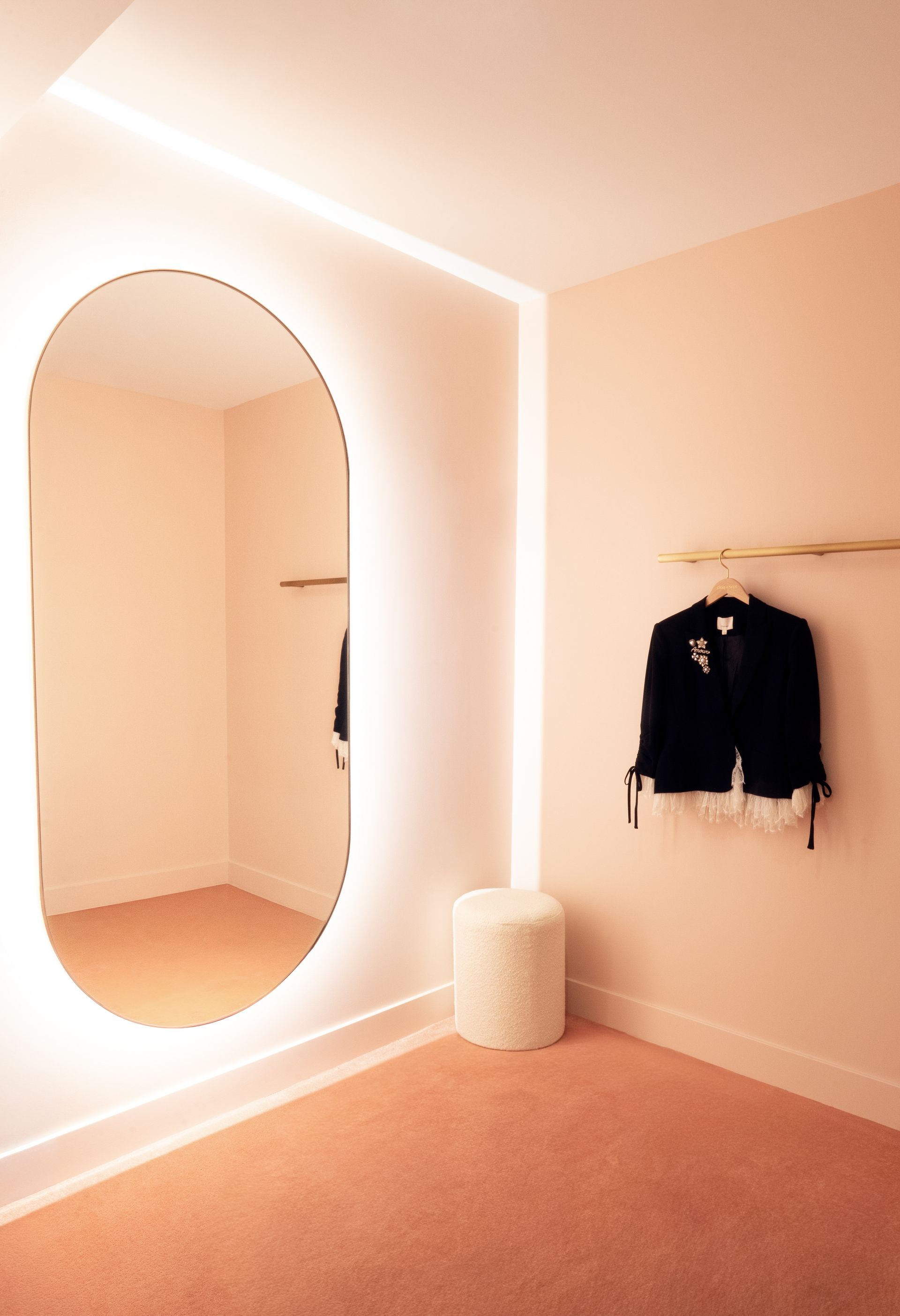Pink fitting room with an oval mirror, warm lighting, and a black jacket hanging on the wall.