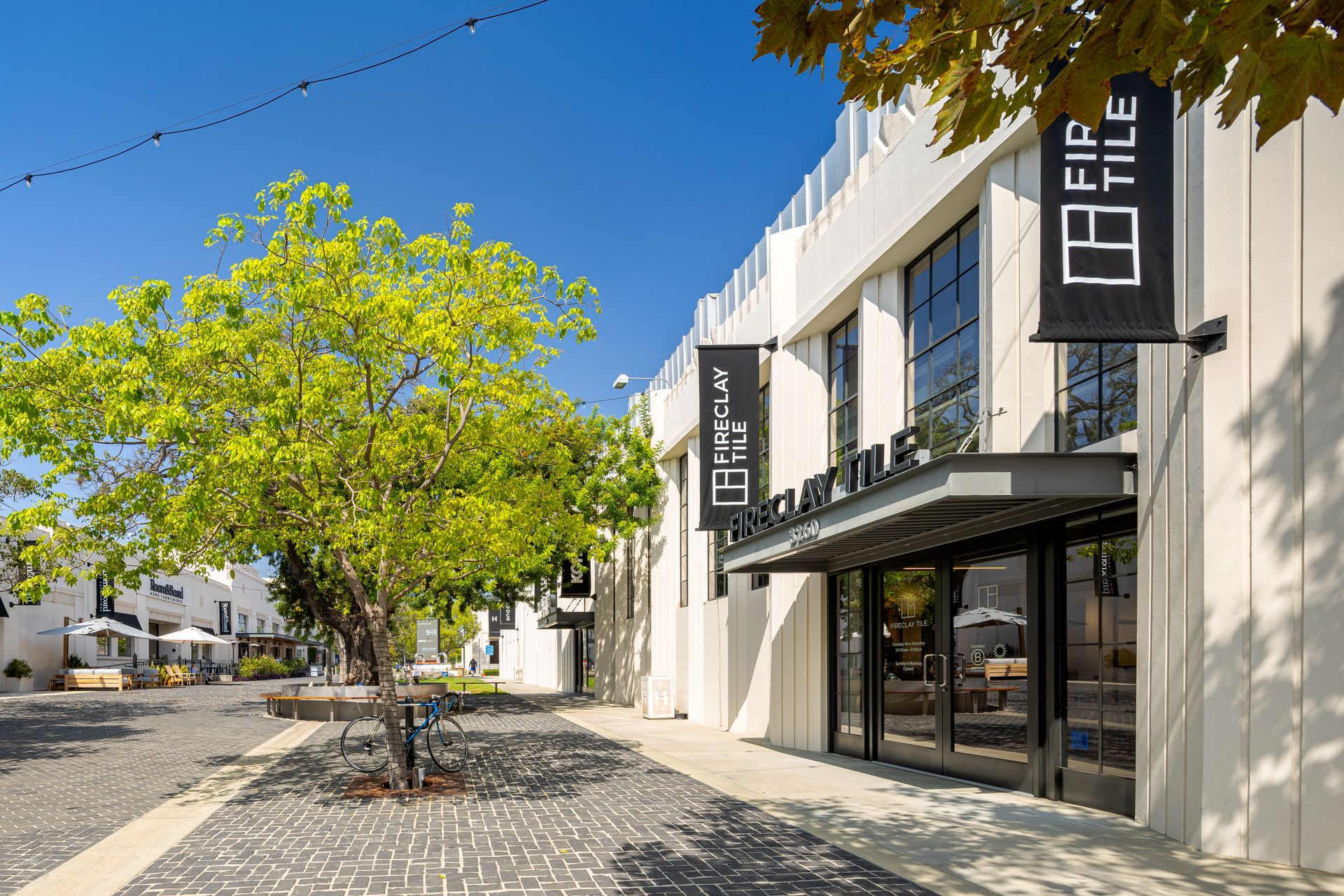 Modern storefront with glass entrance and black signs along a tree-lined cobblestone street