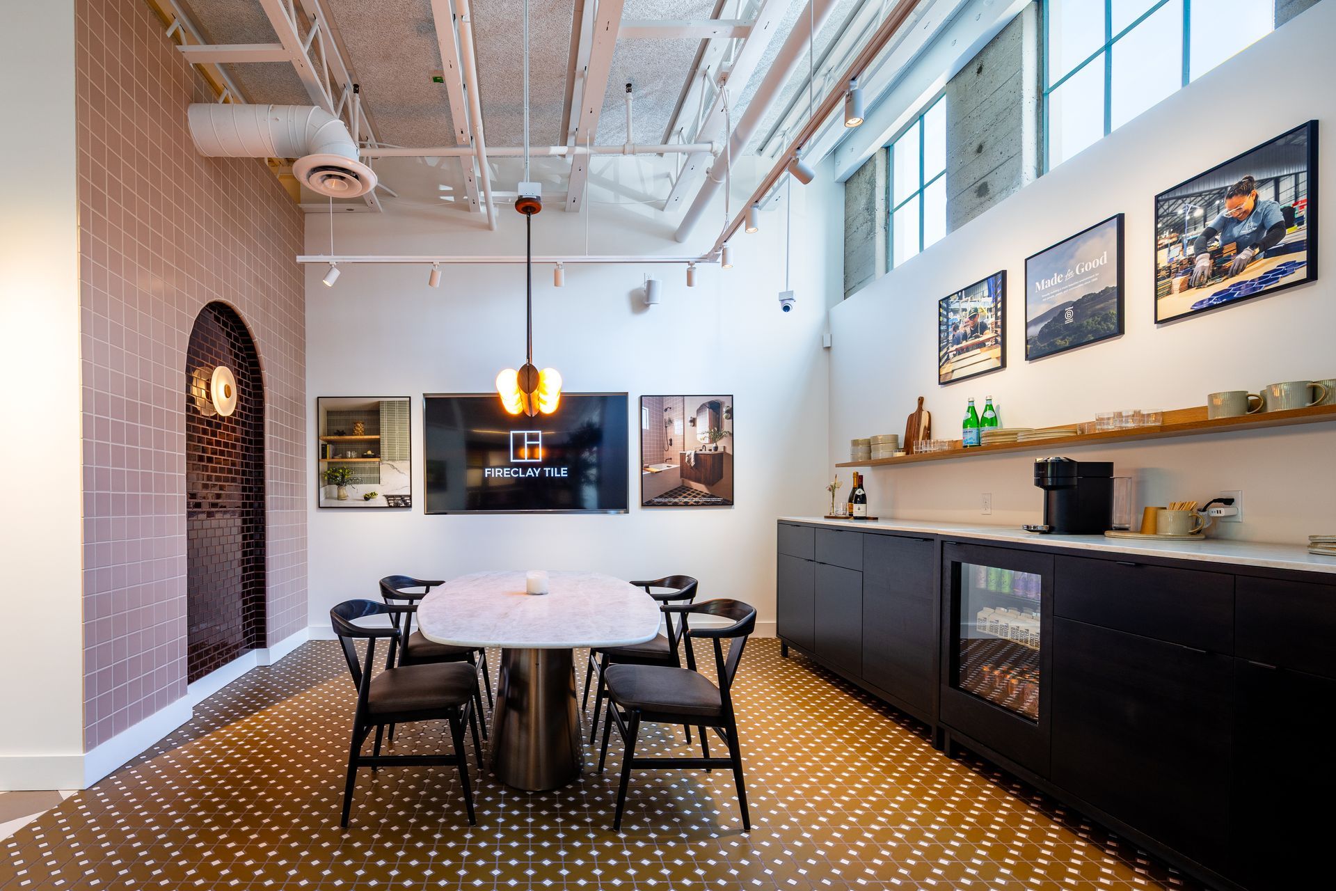 Modern office break room with round table, black chairs, white brick wall, and long counter under framed art.