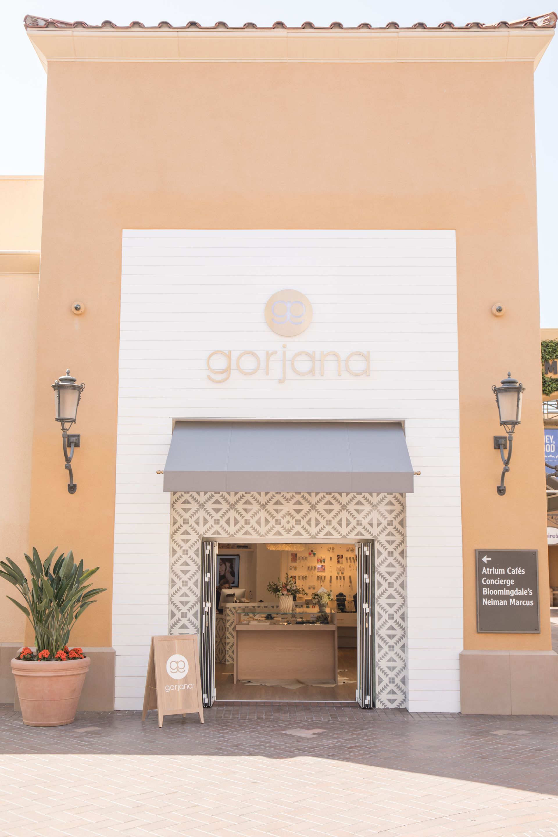 Storefront of a beige shop with a white sign, awning, and open entrance, flanked by potted plants and wall lanterns