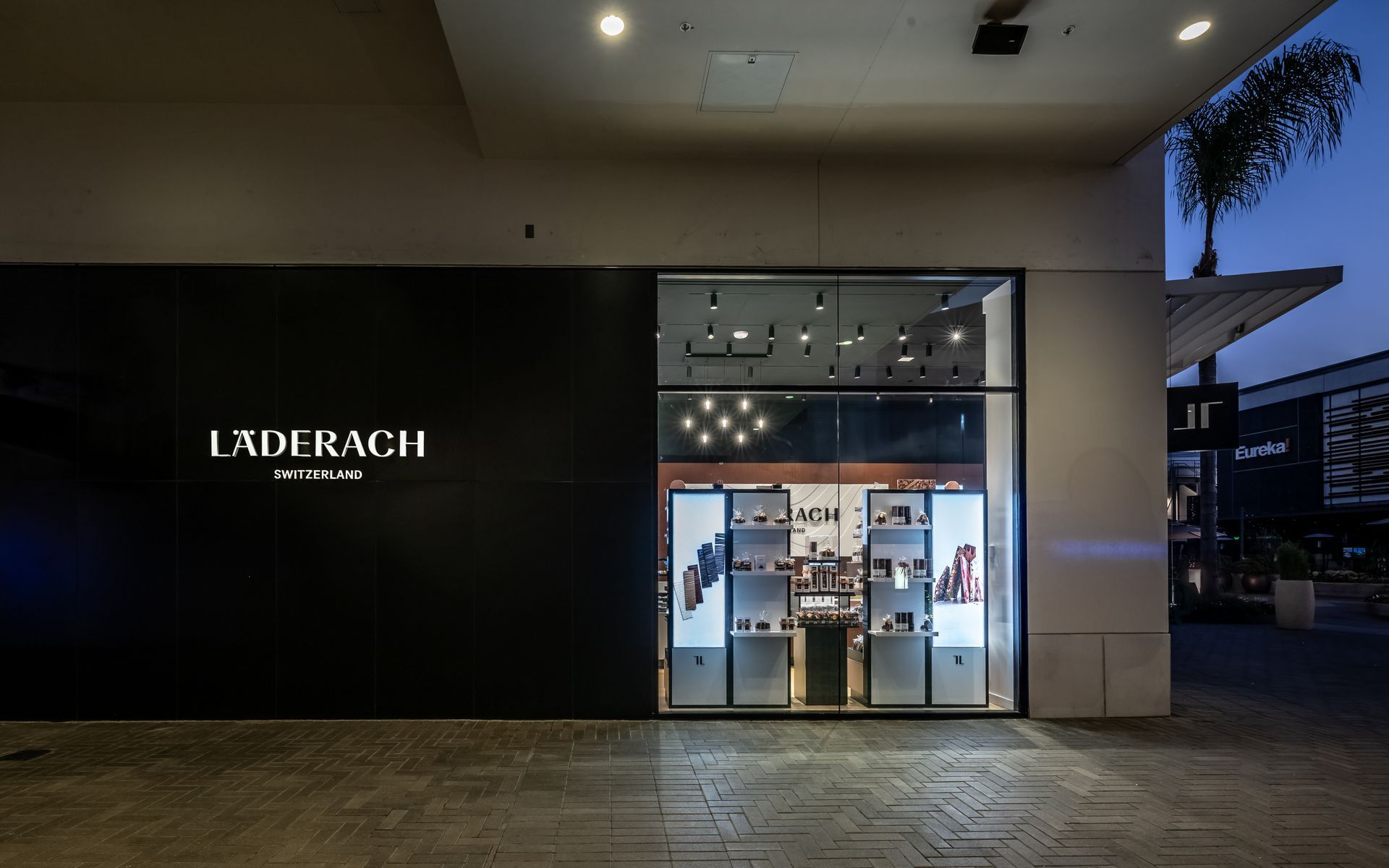 Exterior of an Läderach chocolate shop at dusk, with a lit glass entrance and black storefront.