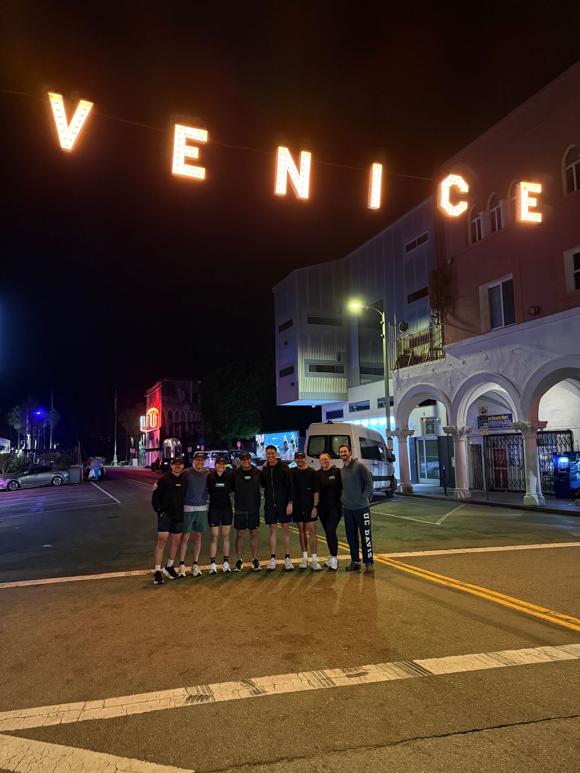 Group of people posing on Venice Beach at night under glowing “VENICE” letters, with boardwalk buildings nearby