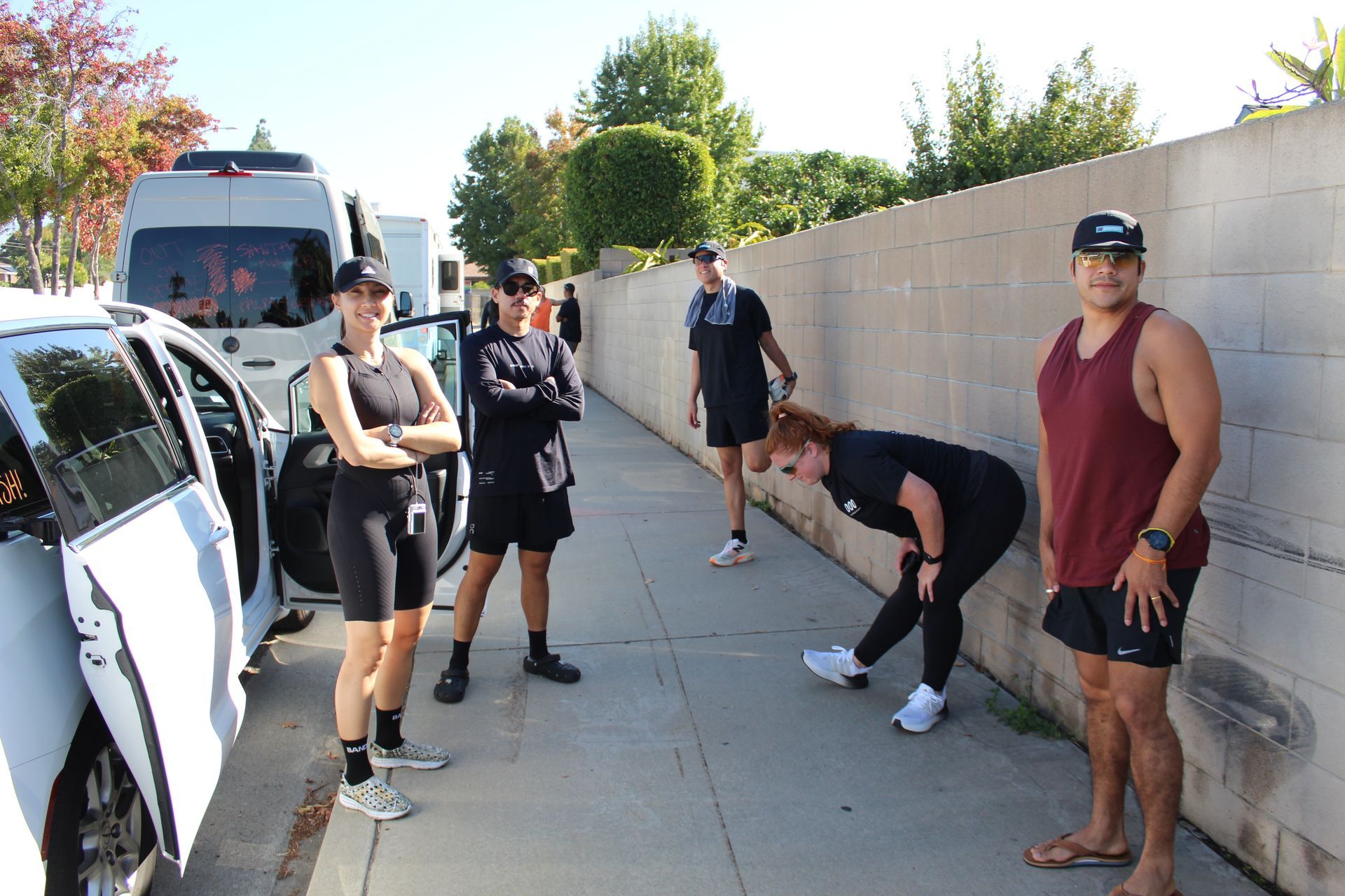 Group of people standing and stretching on a sunny sidewalk beside a wall and parked vehicles