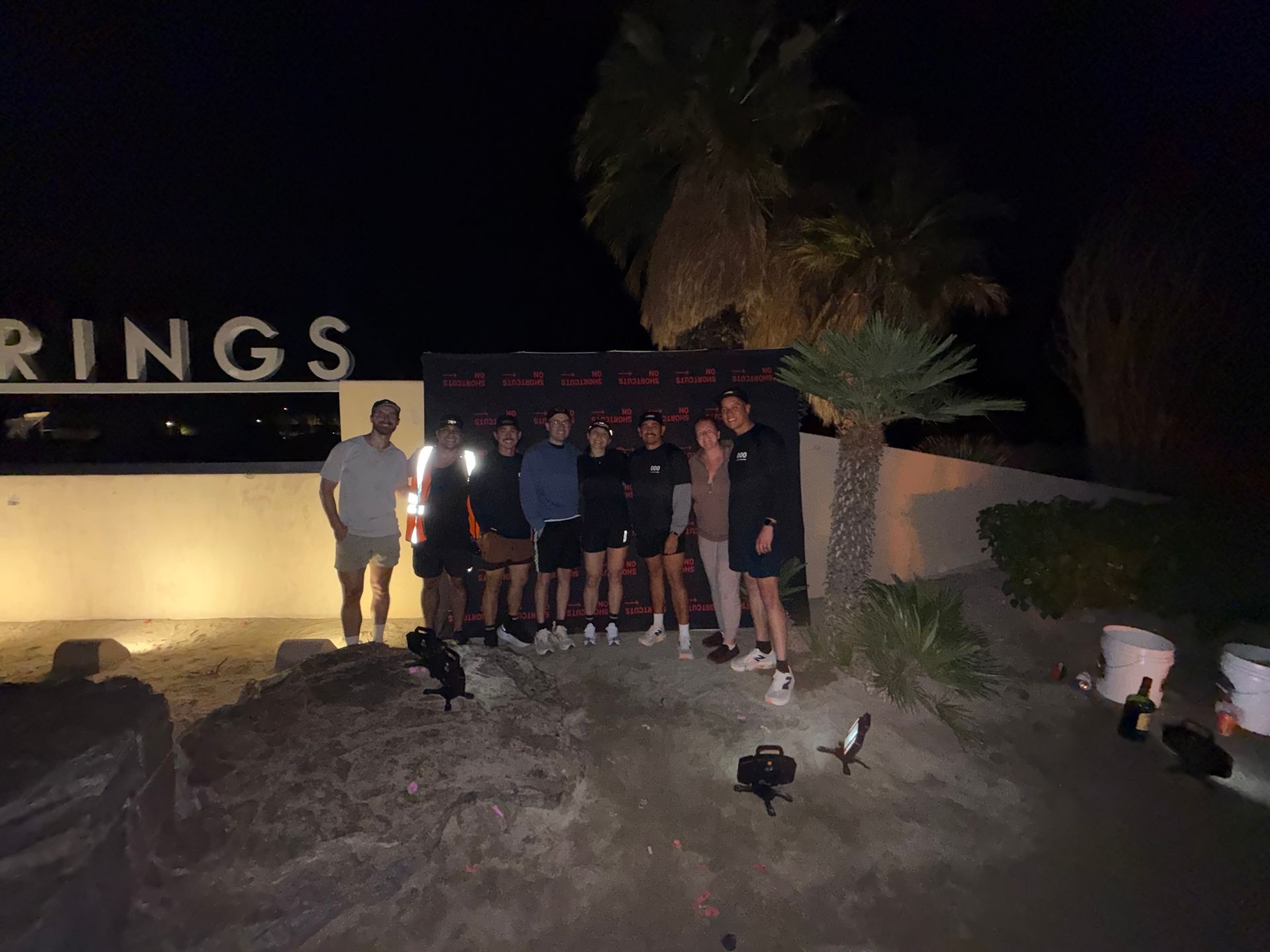Group posing at night beside a lit sign and palm tree, with chickens in the foreground.