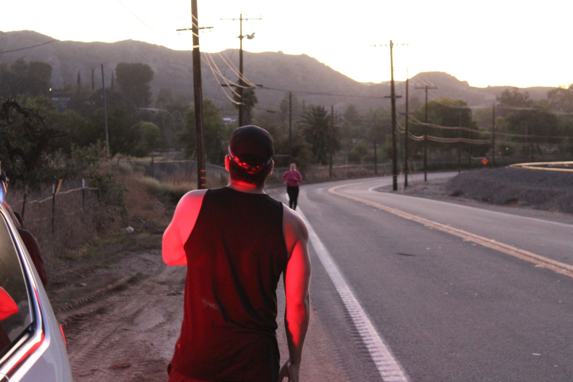 Person in black and red walking on a rural road at sunset with mountains and power lines in the background