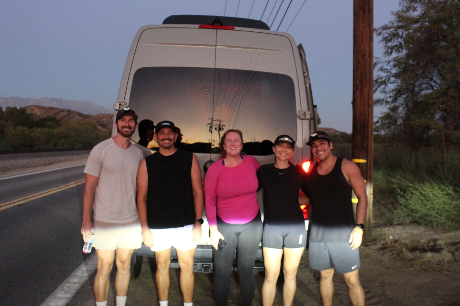 Five people posing in front of a white van on a roadside at dusk