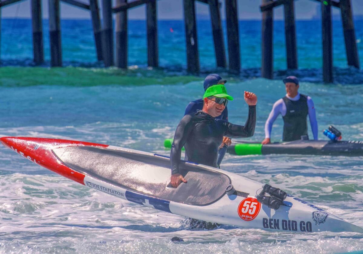 Speedboat racer in black and green gear raises a fist while racing on choppy water near a shoreline
