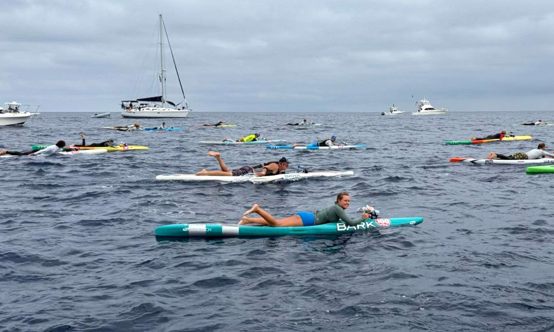 People paddle on surfboards in open water near a sailboat and several swimmers.