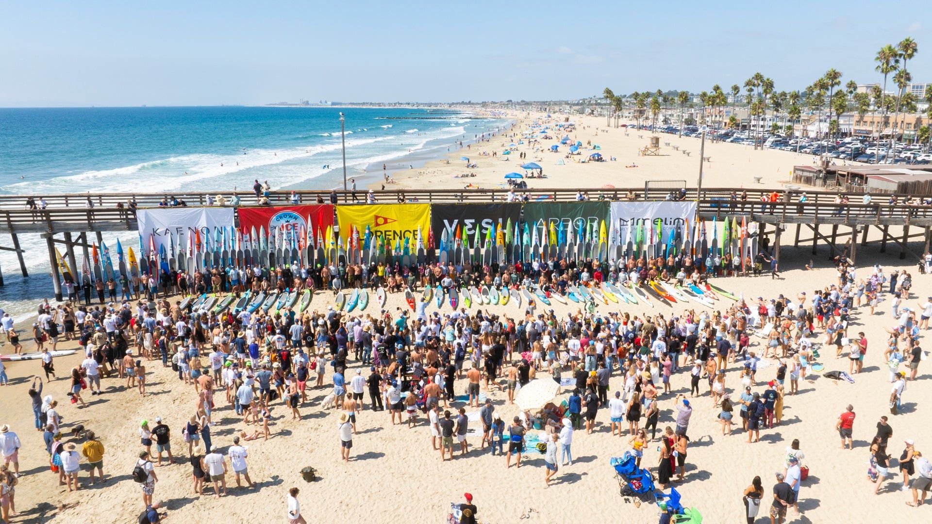 Crowded beach festival with colorful flags, stage, and ocean backdrop on a sunny day