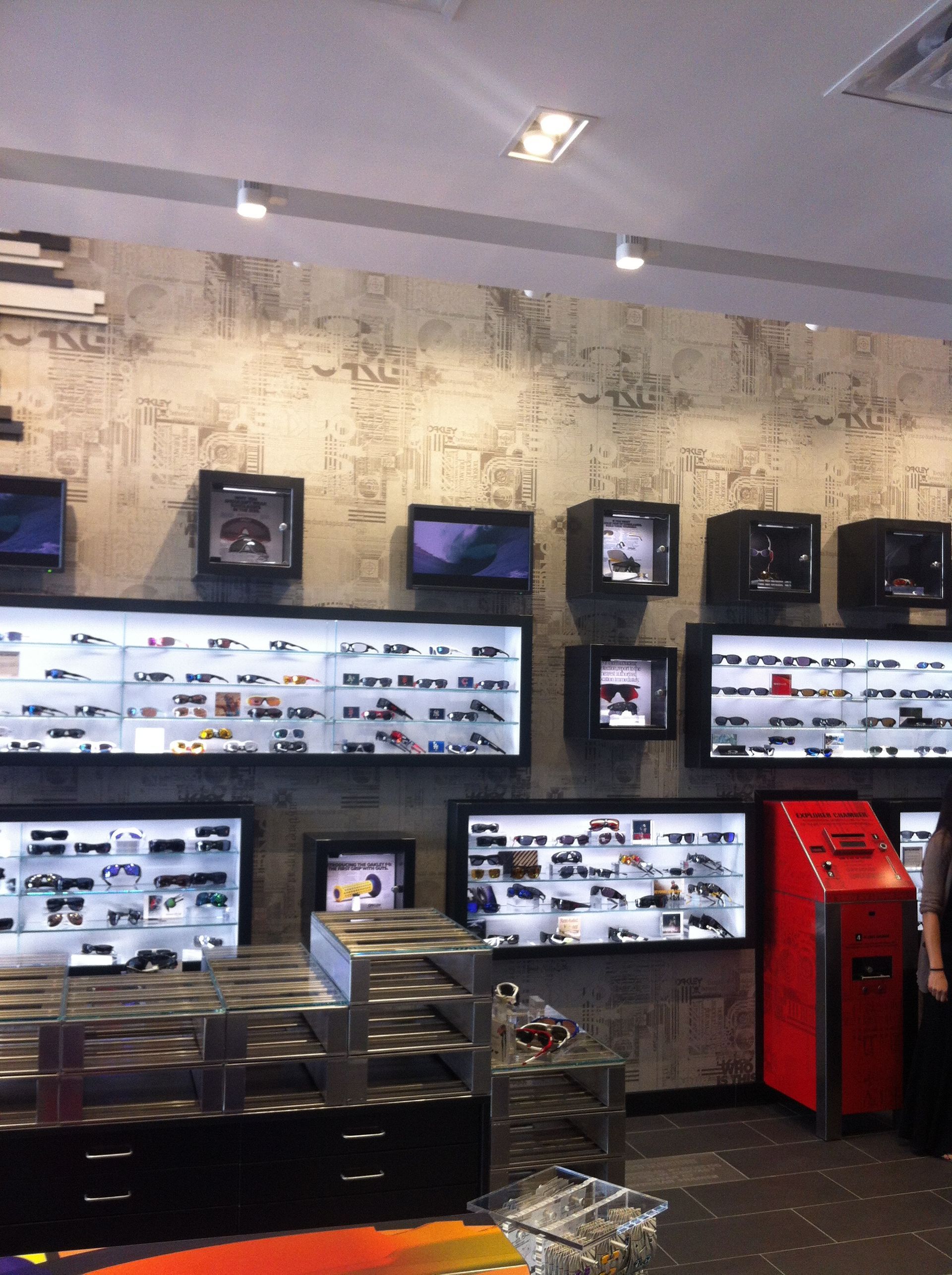 Wall of multiple TV screens and monitors in a modern electronics store, with a red kiosk on the right.