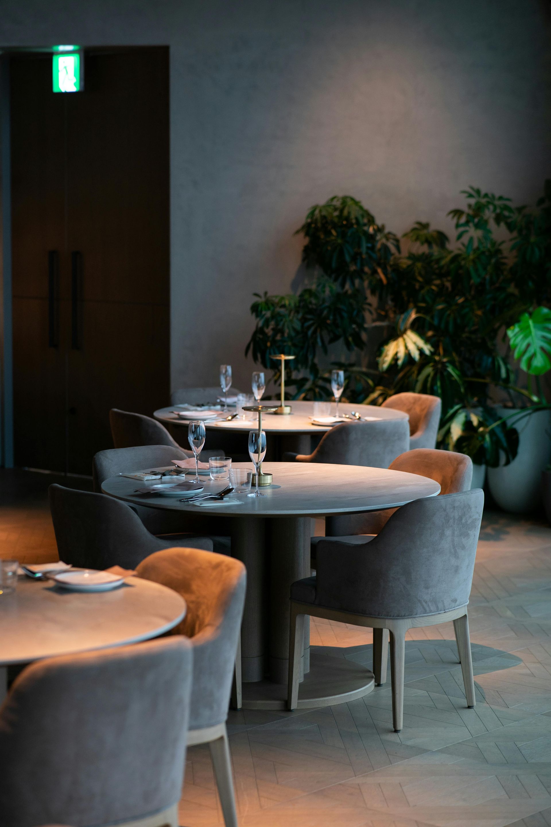 Quiet restaurant dining area with round tables, gray chairs, and potted plants near a wall-lit doorway