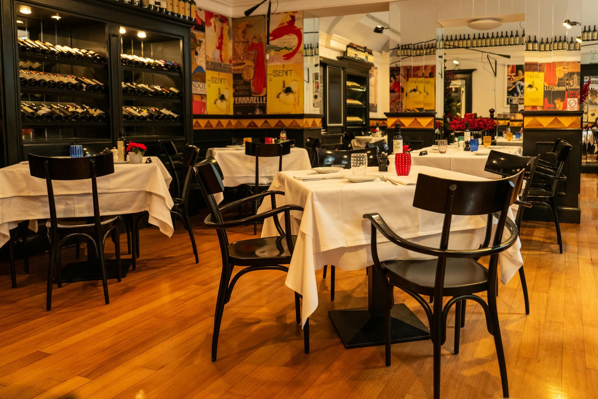 Cozy restaurant dining room with white tablecloths, black chairs, wine racks, and warm wood floors.
